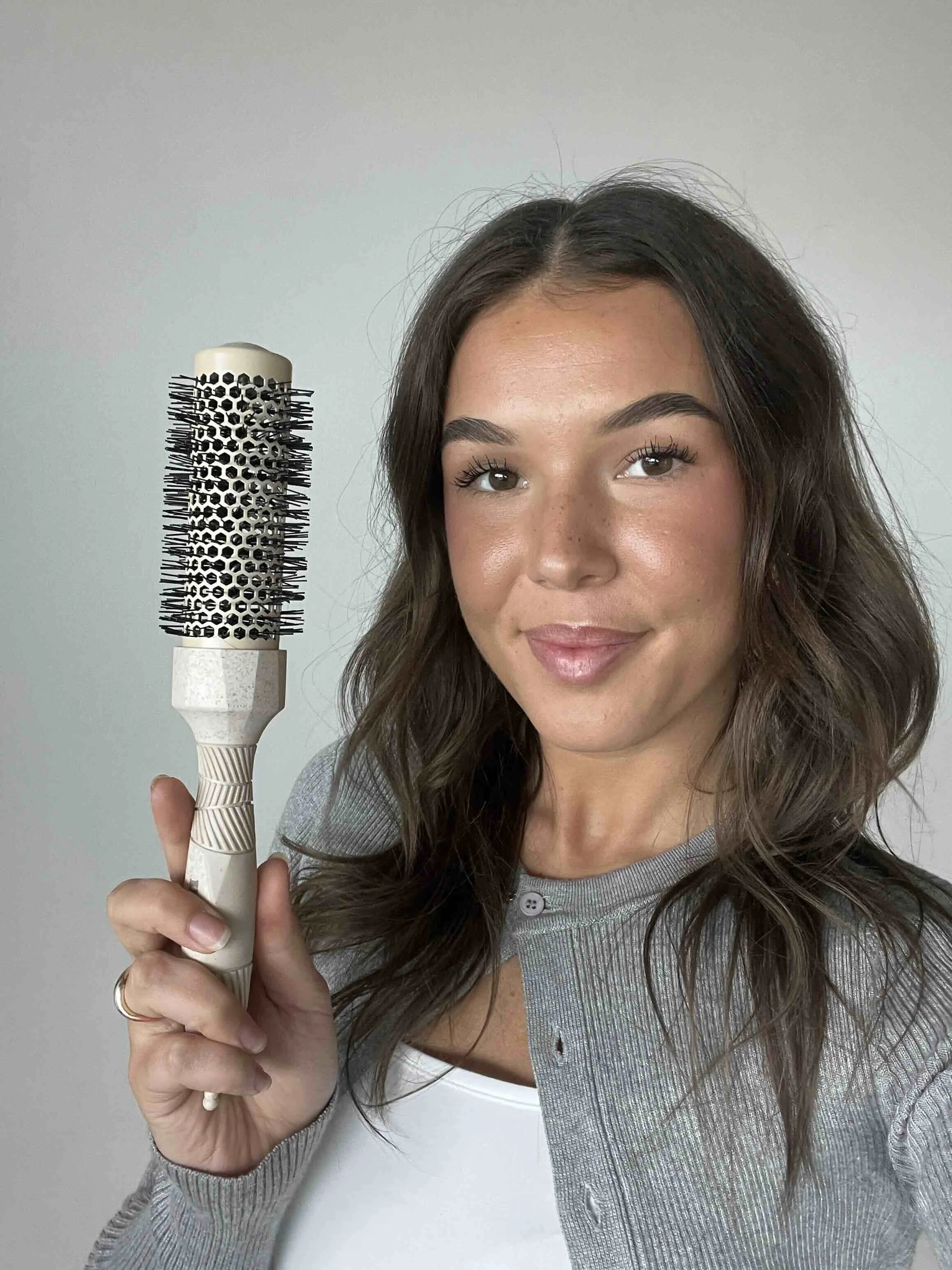Woman holding a round hairbrush with a wooden handle, wearing a gray cardigan over a white top, standing against a plain background.