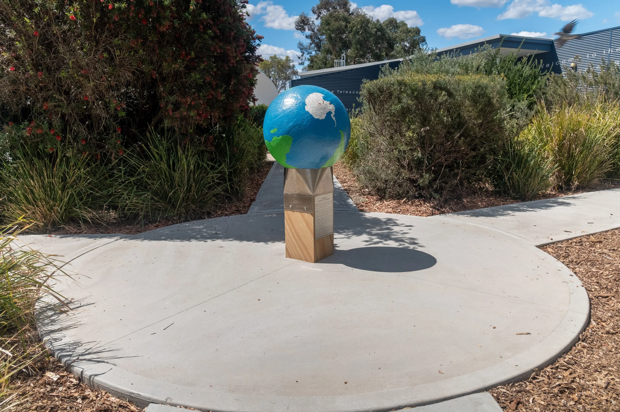 A painted globe sculpture on a pedestal, situated on a circular concrete platform outdoors with bushes and trees in the background.