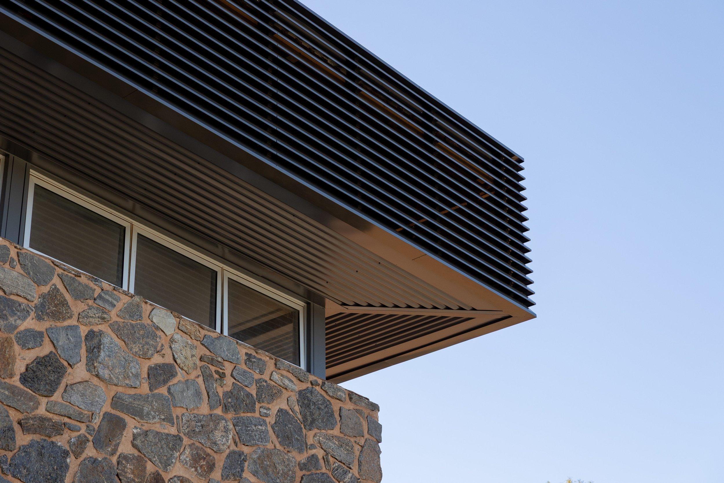 Close-up of a modern building's upper corner, featuring a stone wall, large window, and metal louvers under a clear blue sky.