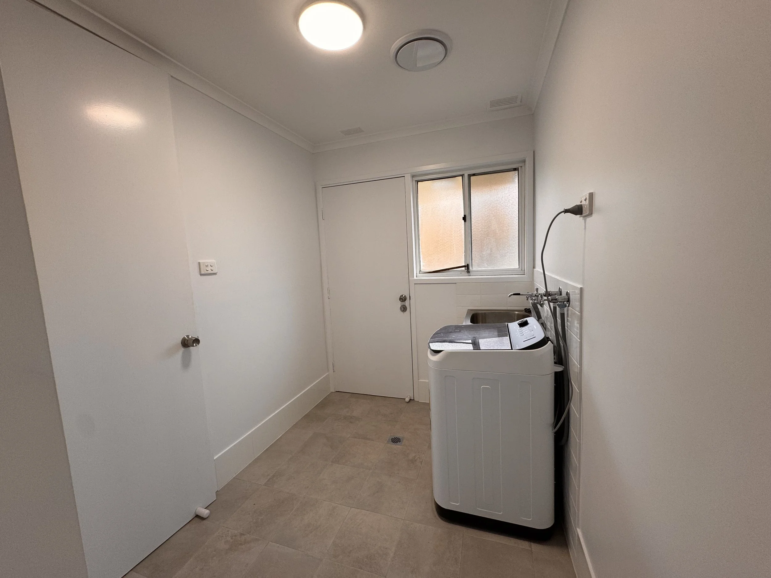 Laundry room with white walls, beige tile floor, a window, a white washing machine, and a small stainless steel sink.