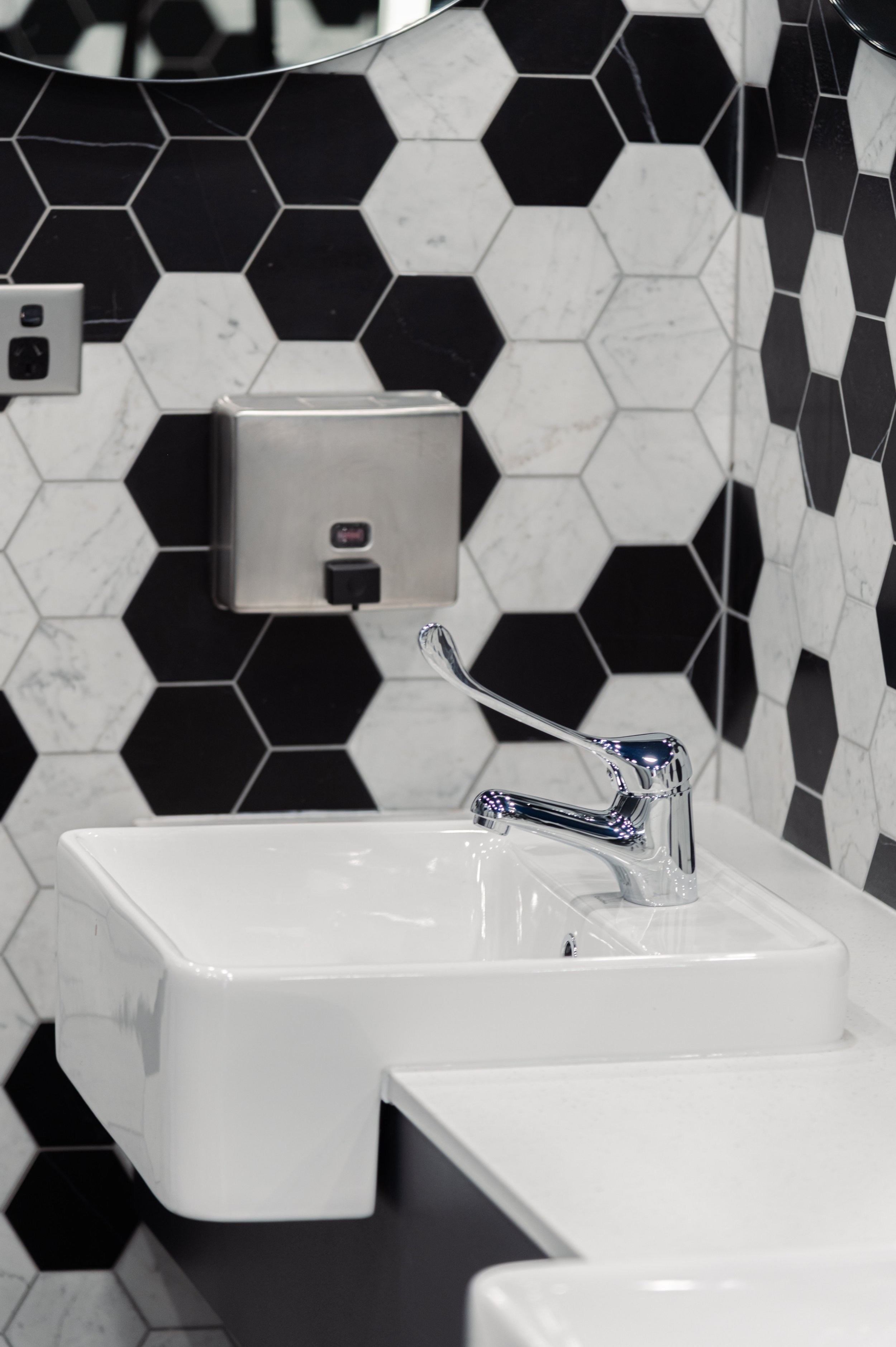 A small white wall-mounted sink with a chrome faucet, black and white hexagonal tile wall, and a stainless steel hand sanitizer dispenser.