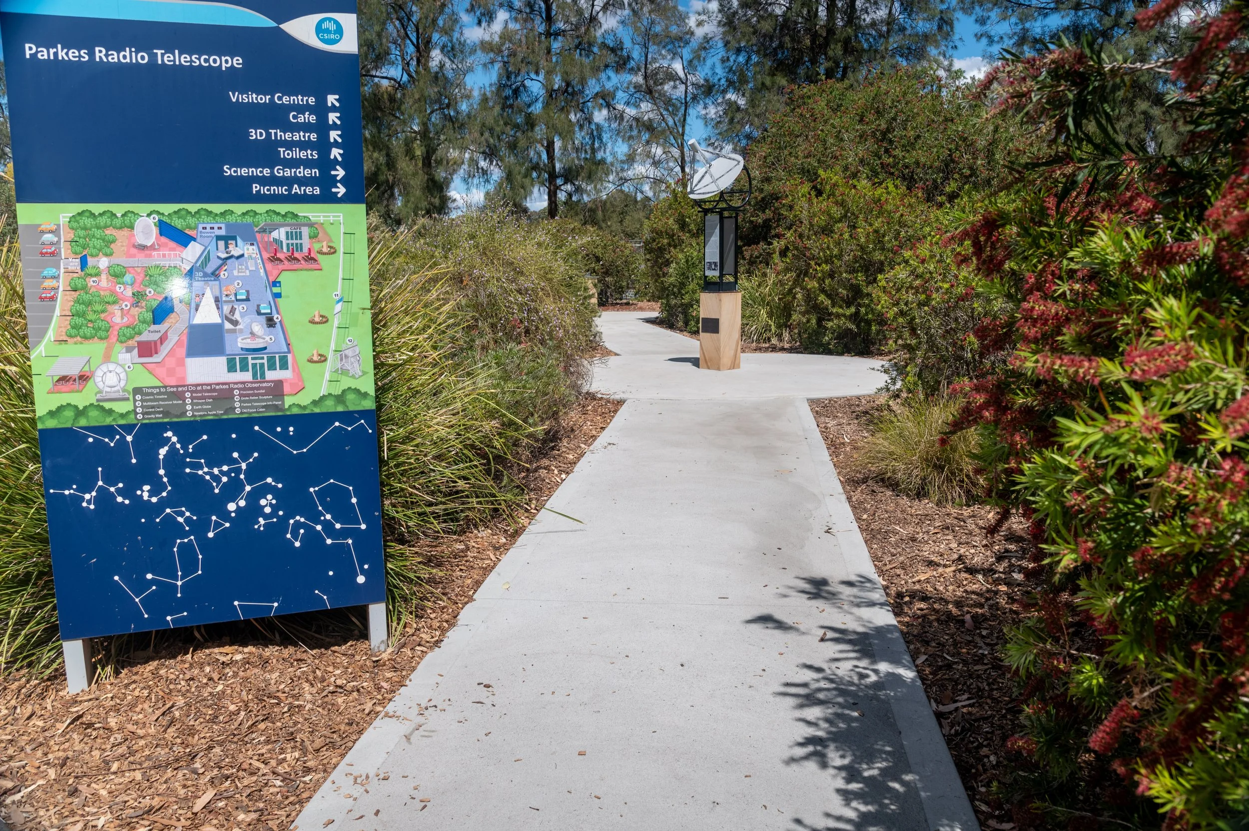 Walkway leading to the Parkes Radio Telescope with a colorful map sign on the left and a satellite dish on the right, surrounded by bushes and trees.