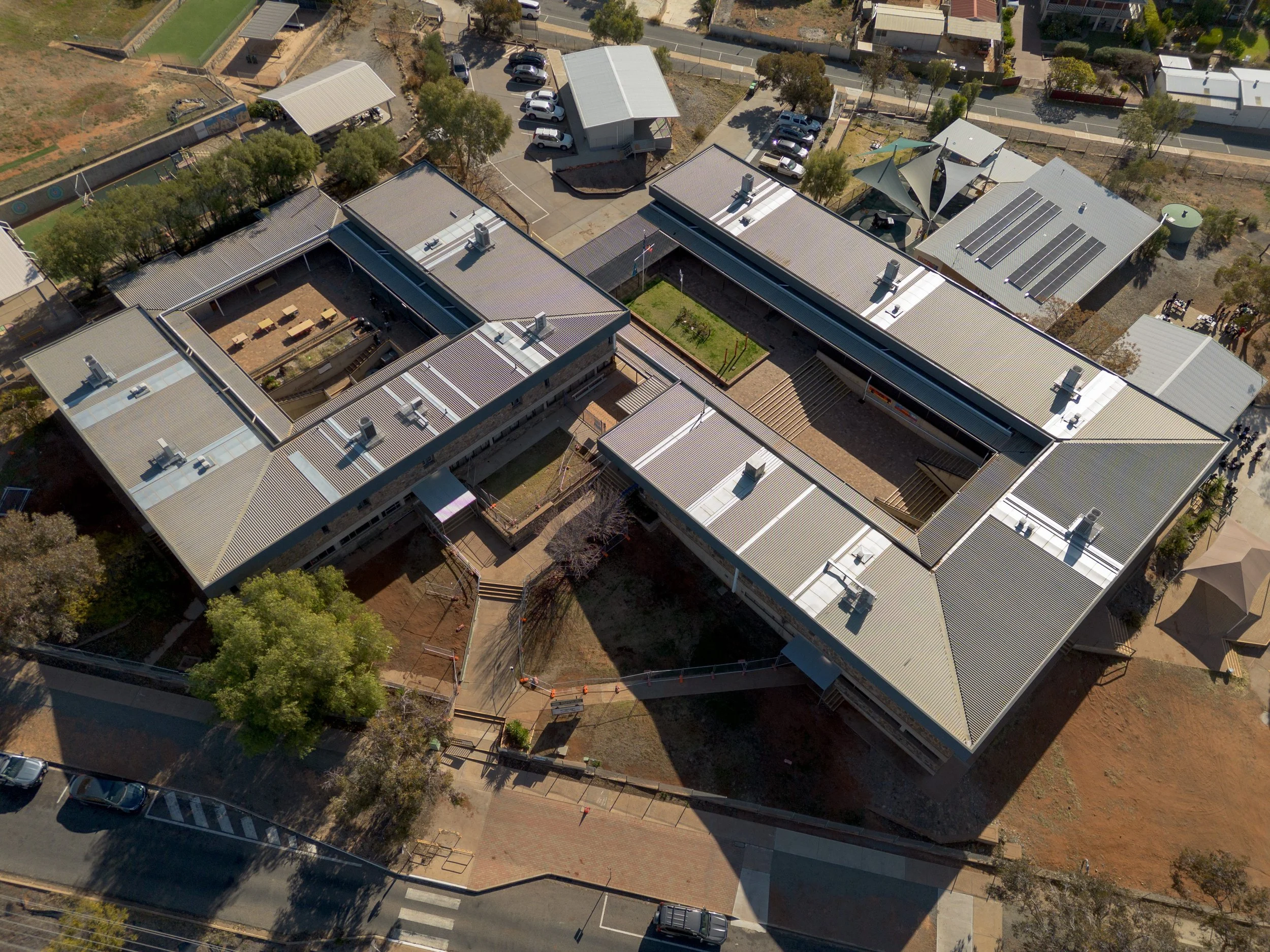 Aerial view of a school campus with multiple single-story buildings, outdoor courtyard, parking lot, and surrounding trees.