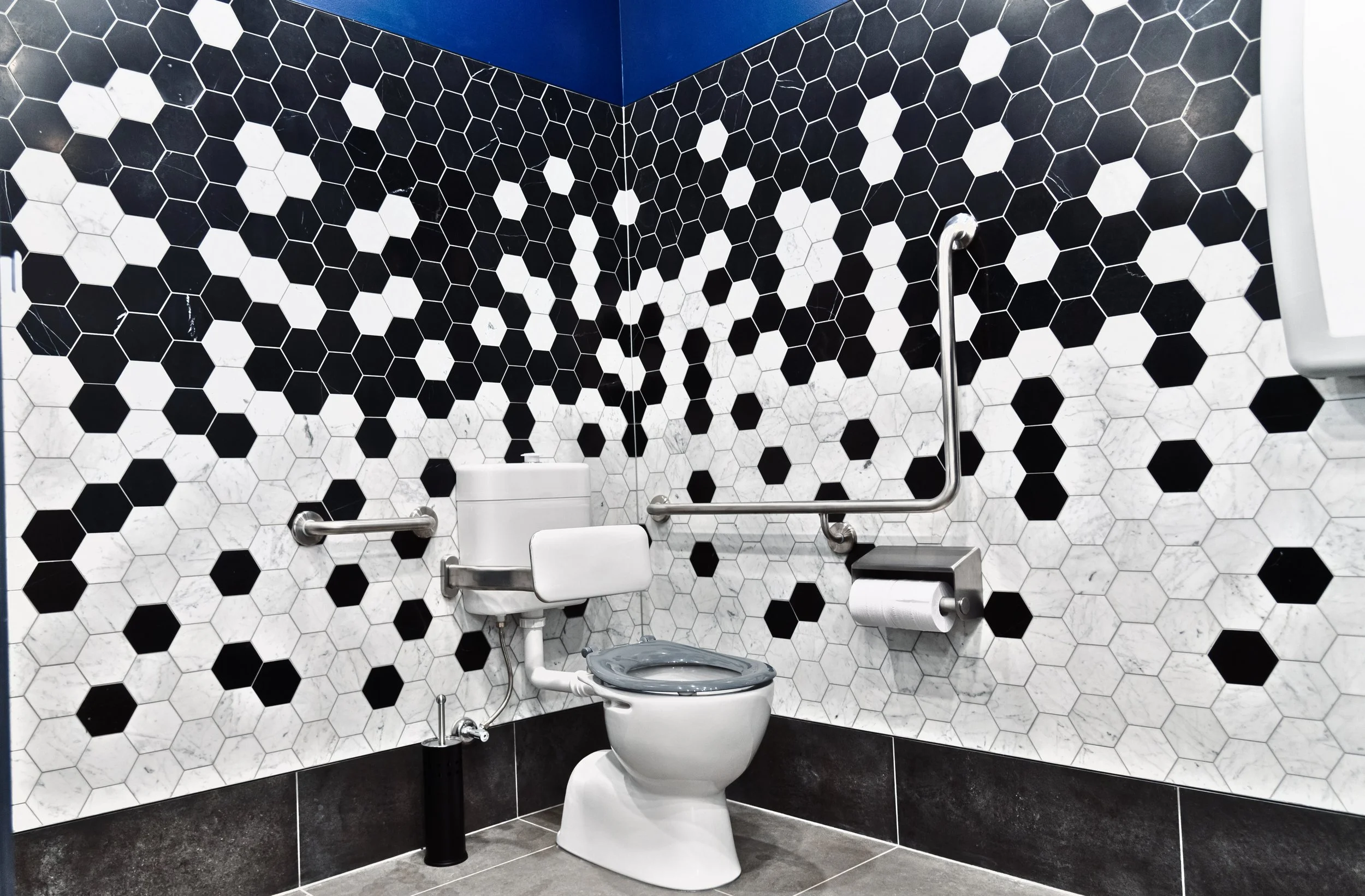 Accessible restroom with black and white hexagonal tile walls, a white toilet with a gray seat, and metal grab bars on the walls, with a roll of toilet paper on a holder.