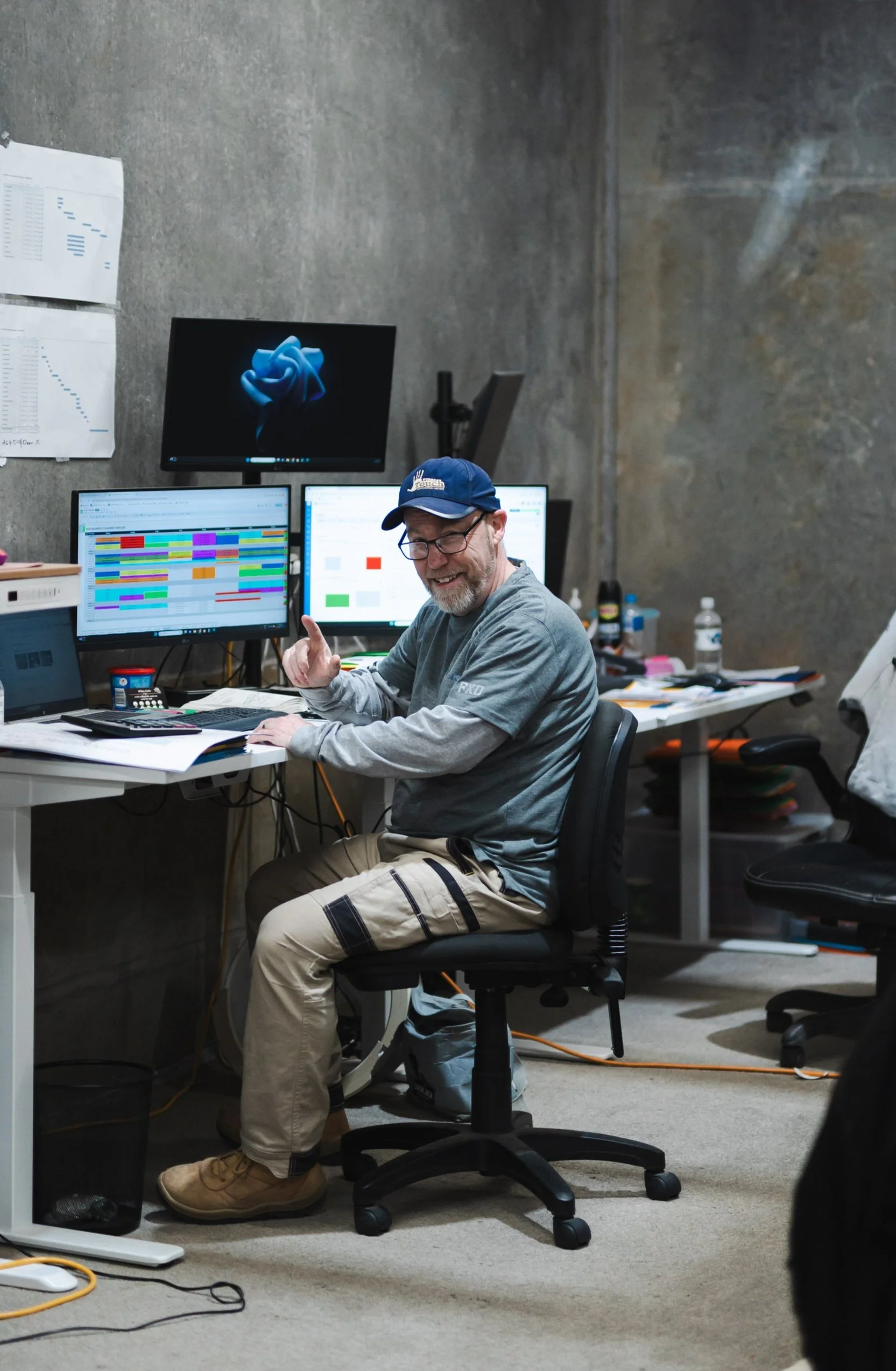 A man with glasses, a beard, and a blue cap, smiling and giving a thumbs-up, sits at an office desk with multiple monitors displaying spreadsheets and charts in a cluttered office.