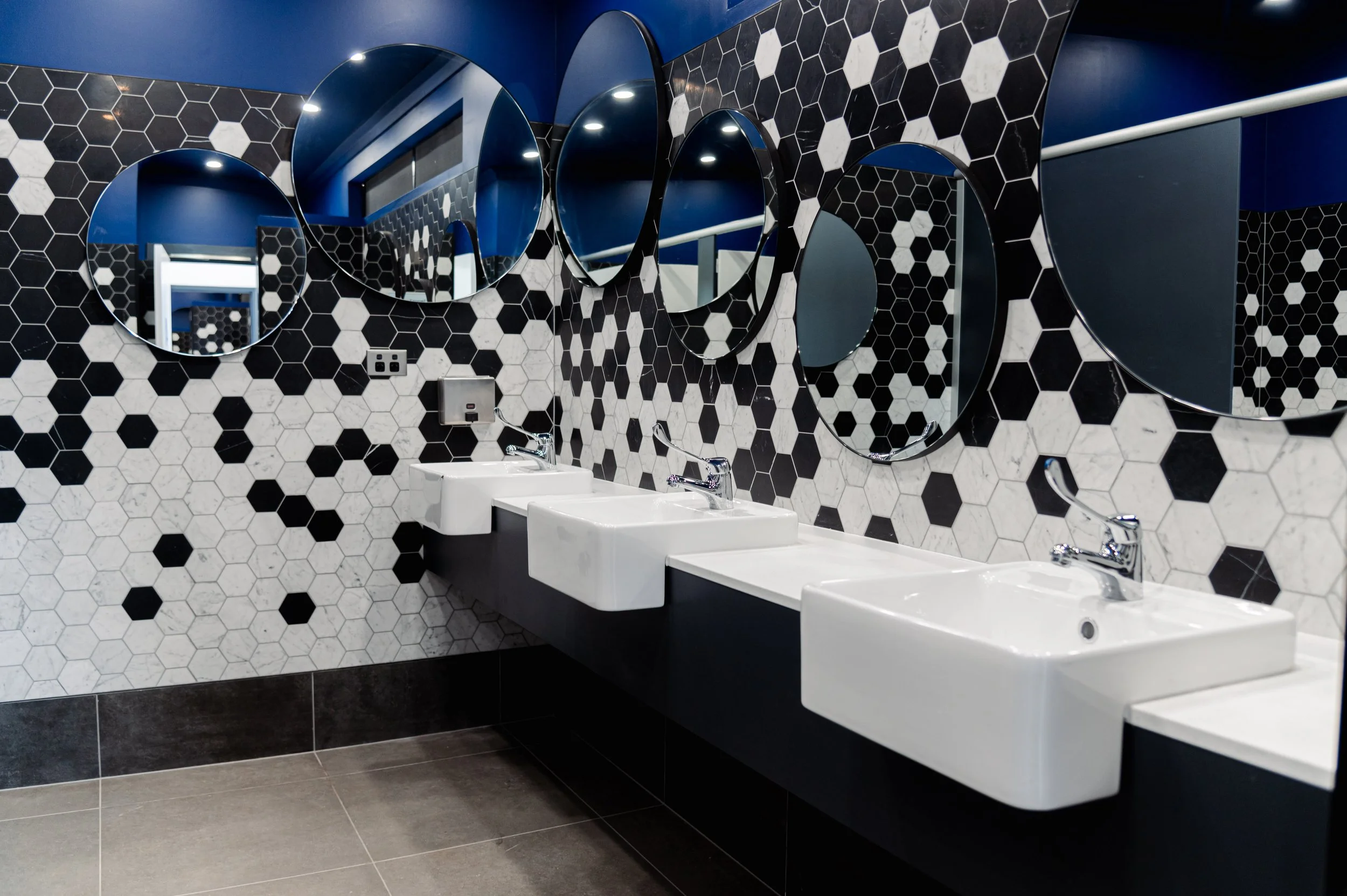 A modern public restroom with four white sinks mounted on a black counter, a black and white hexagon patterned tiled wall, three circular mirrors, and a blue ceiling.