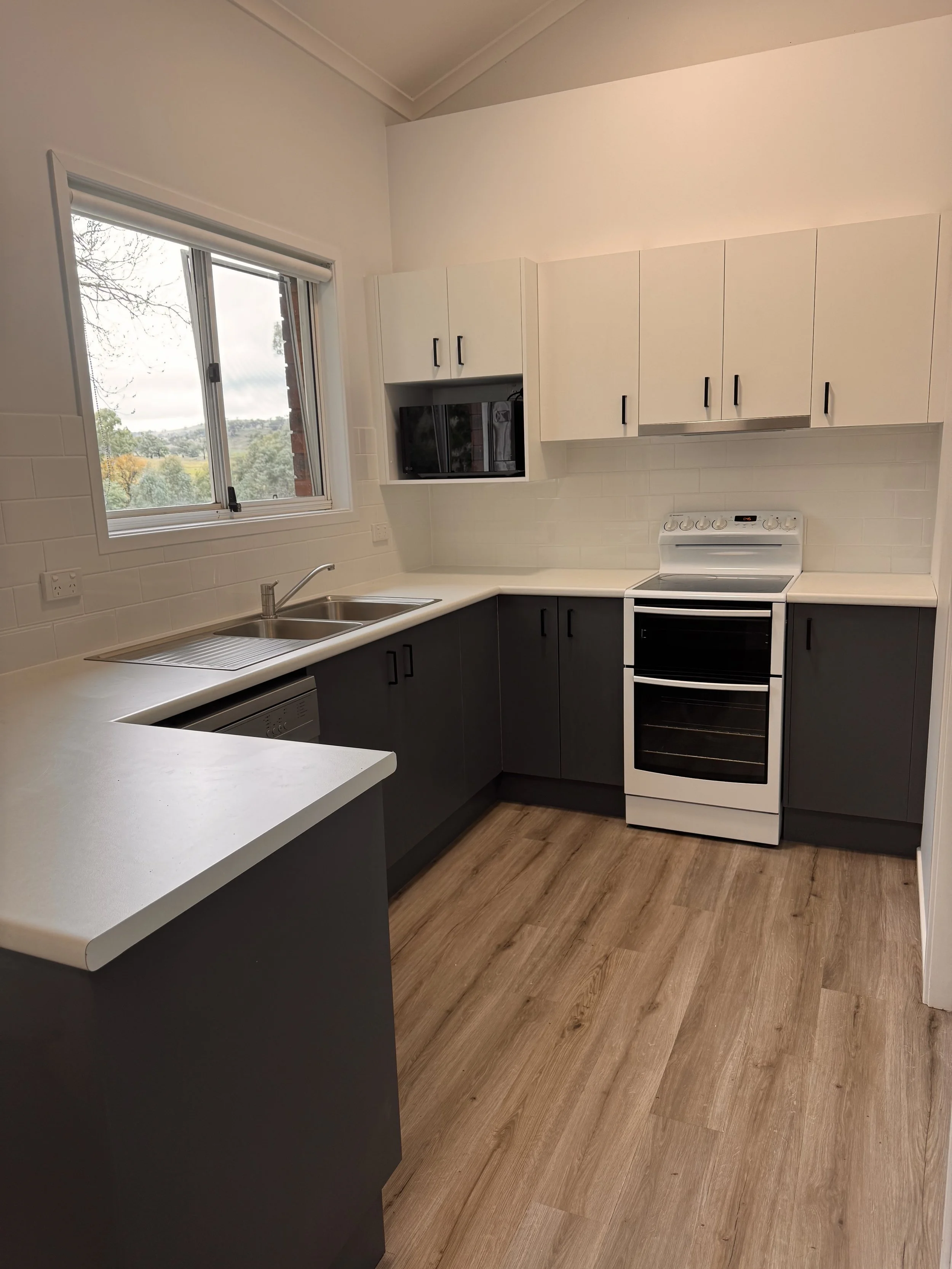 Modern kitchen with white upper cabinets, dark lower cabinets, white countertops, a stainless steel sink, a built-in microwave, a white stove with oven, a window above the sink, and wood-look flooring.