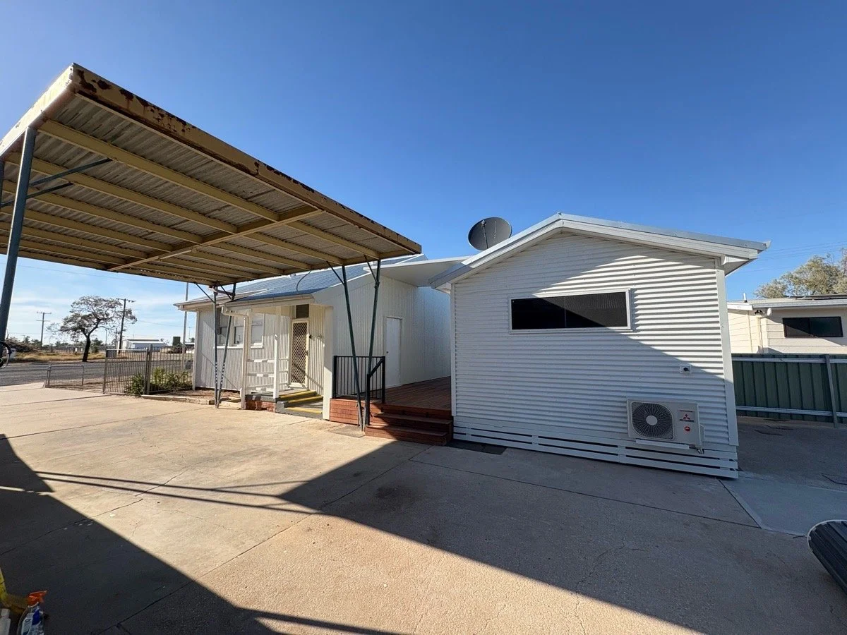 A white building with siding, a black window, and a satellite dish on the roof, with a large metal carport attached, located on a concrete lot under a clear blue sky.