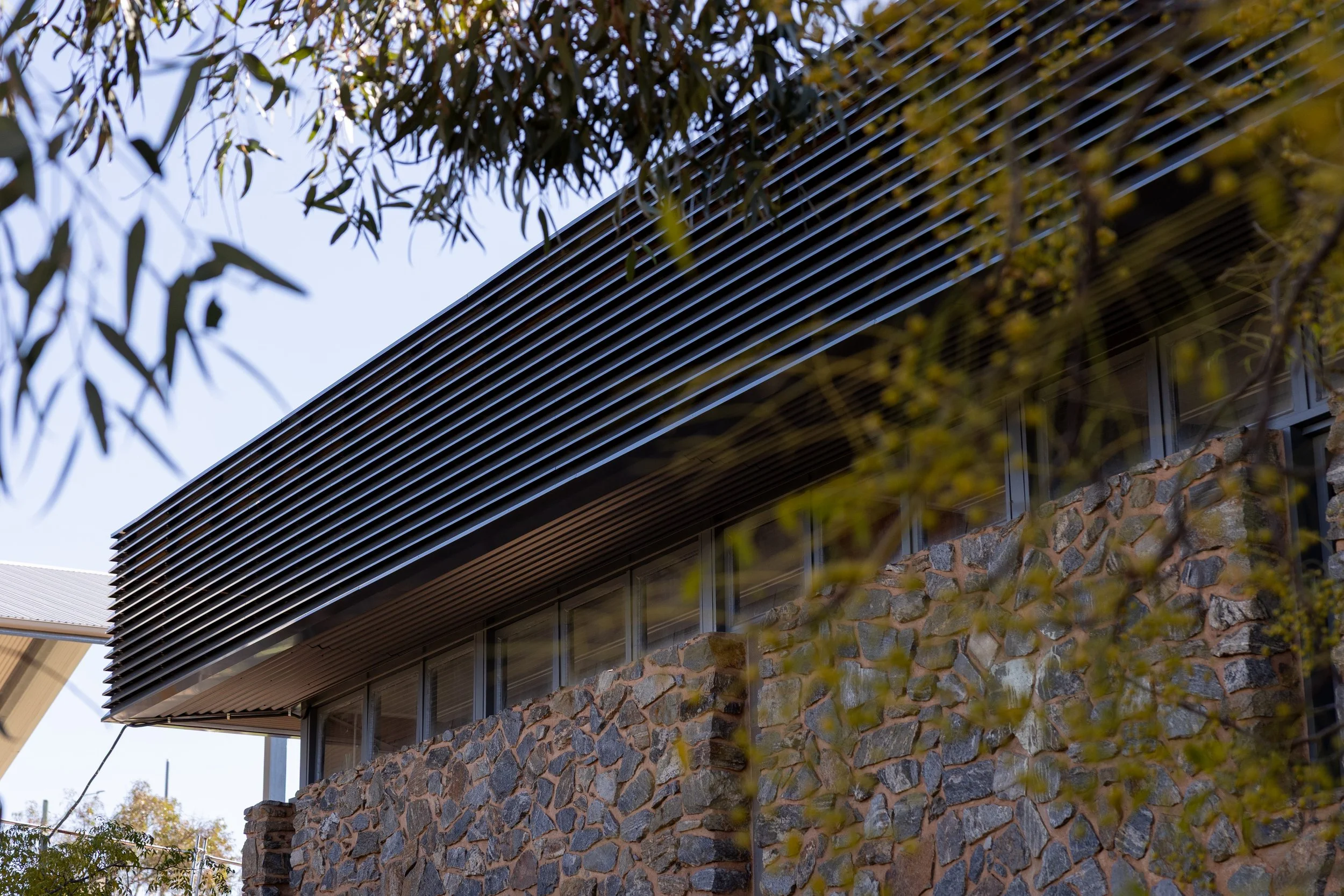 Close-up of a modern building with stone exterior and black metal slatted overhang, partially obscured by tree branches.