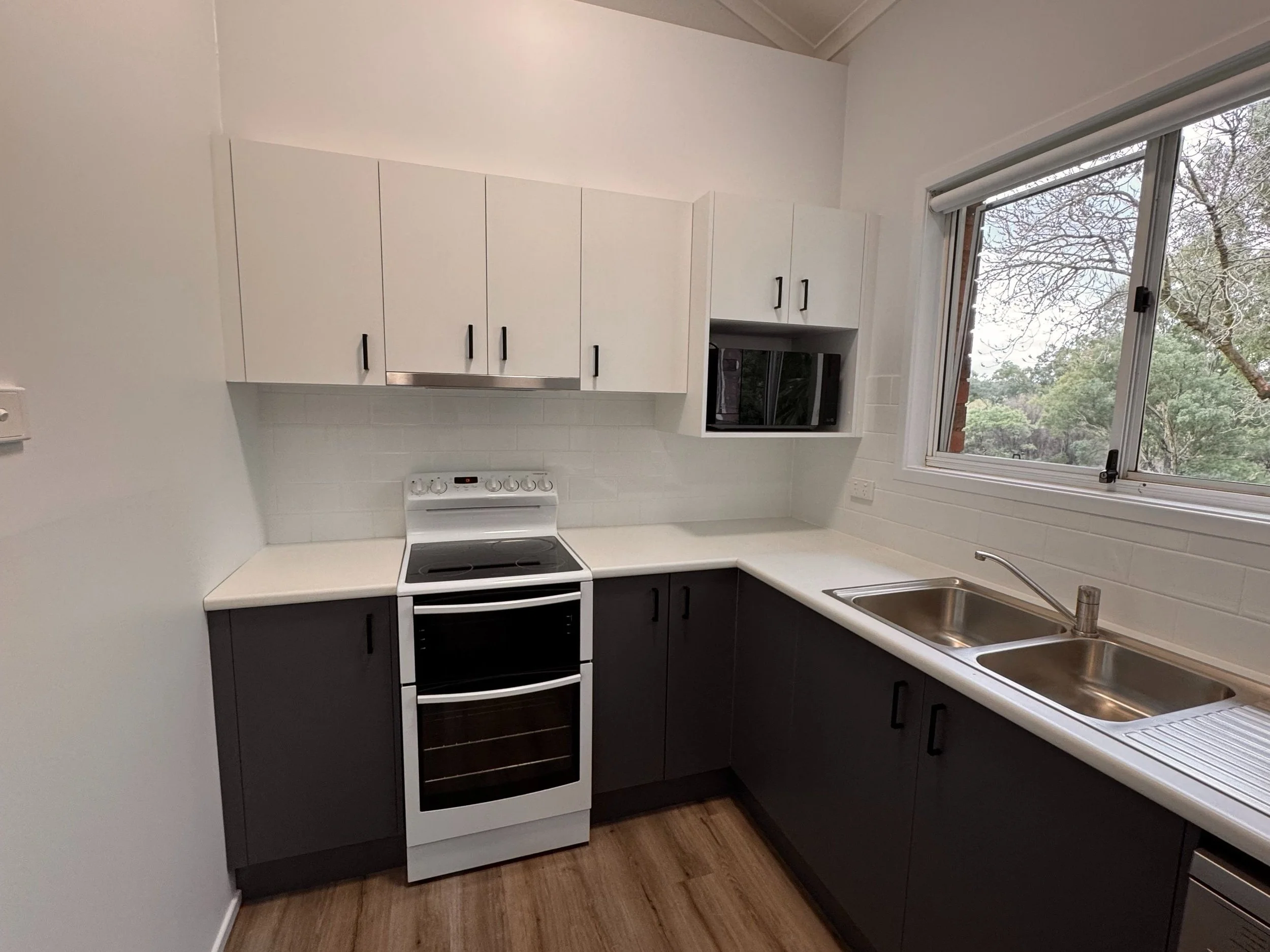Kitchen with white upper cabinets, dark lower cabinets, white countertop, double sink, stove, and microwave, with a window showing trees outside.