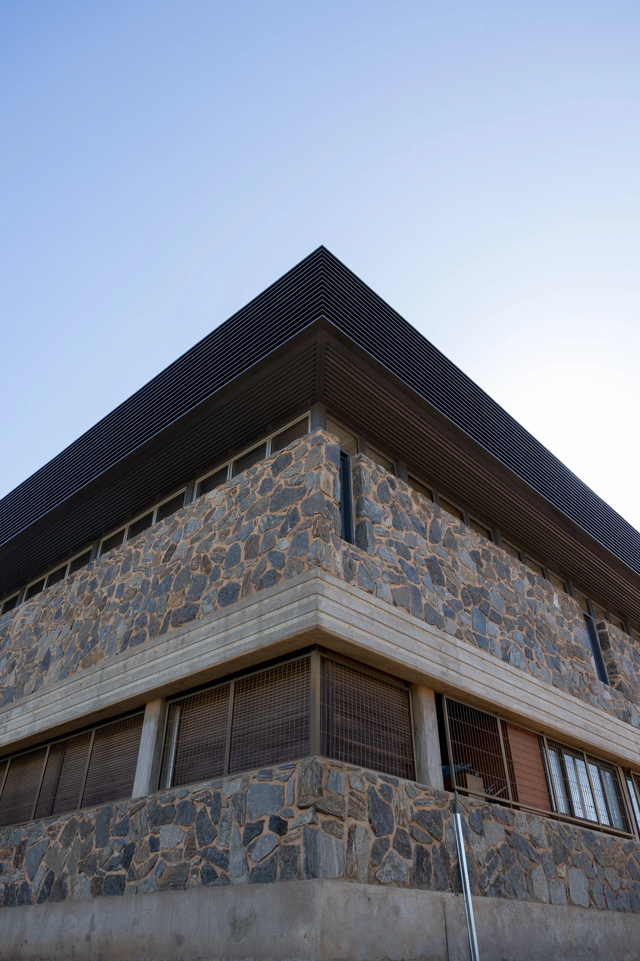 Close-up of a modern stone and concrete building corner with windows and black metal roofing against a clear blue sky.