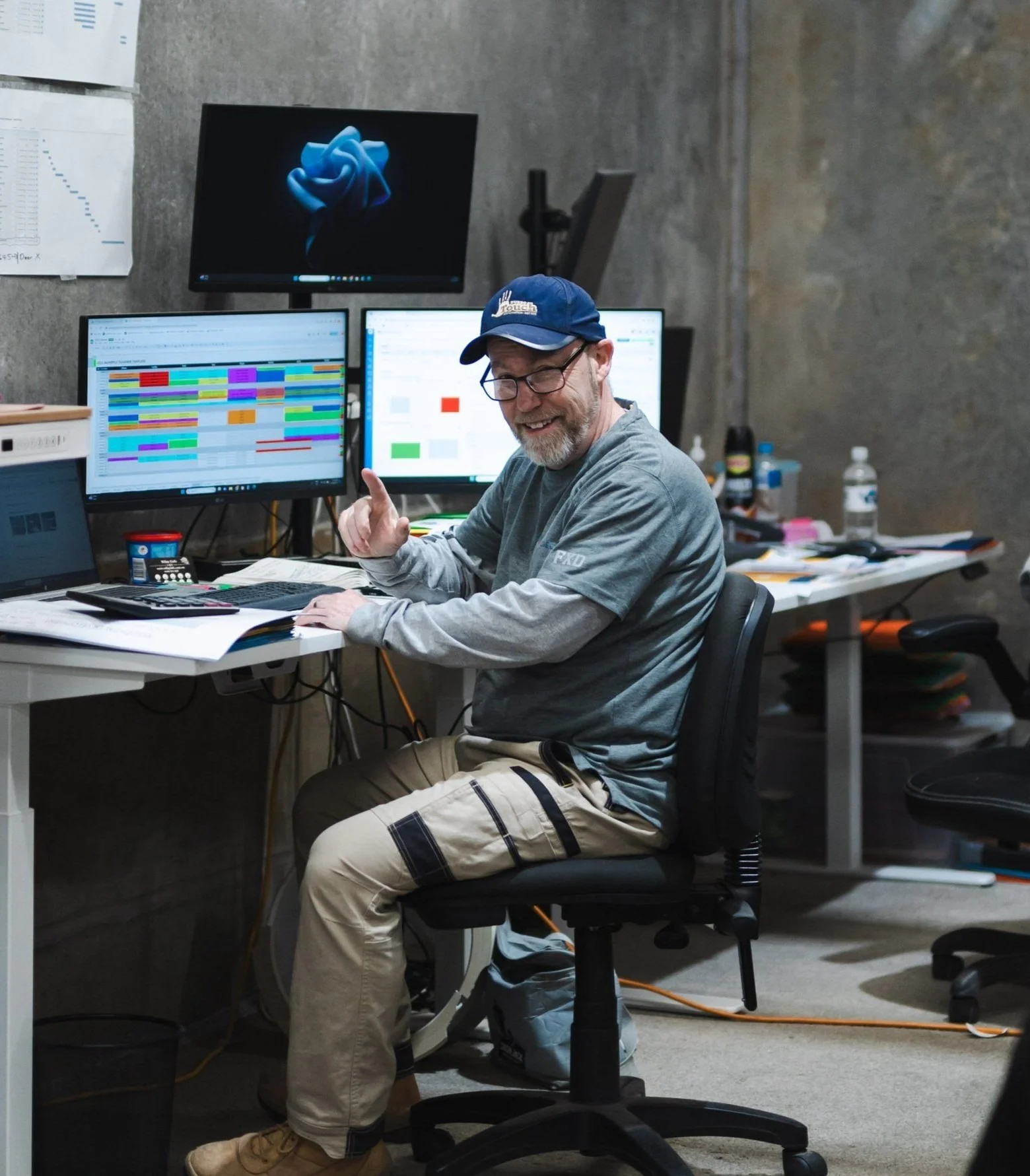 Man sitting at a cluttered desk in an office, smiling and pointing, with multiple computer monitors displaying colorful spreadsheets or charts.