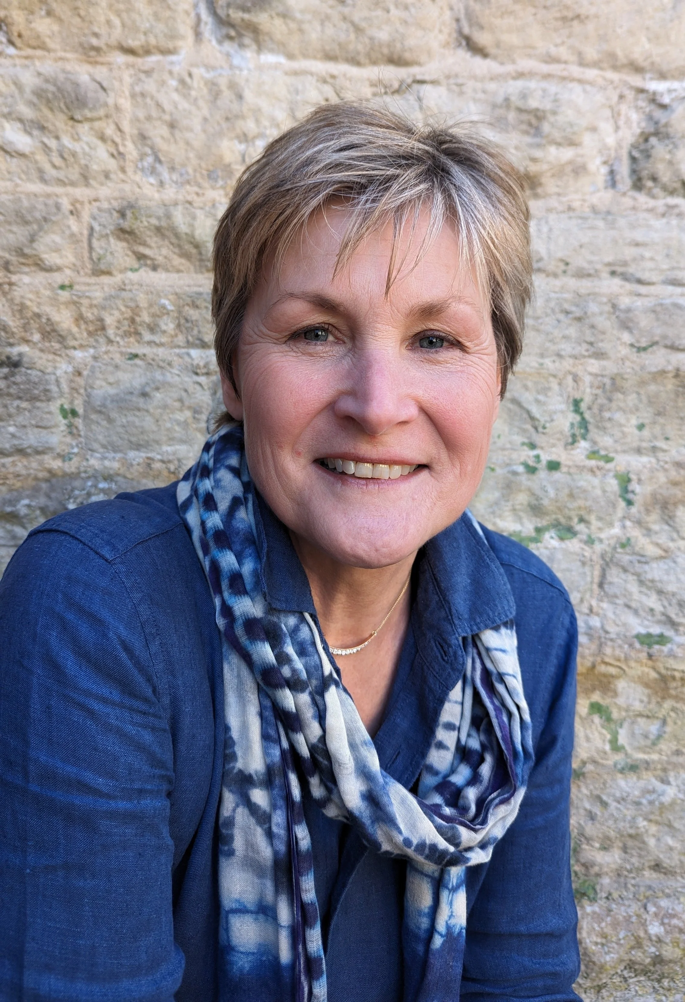 A middle-aged woman with short blonde hair, wearing a blue shirt and a patterned scarf, smiling in front of a stone wall.