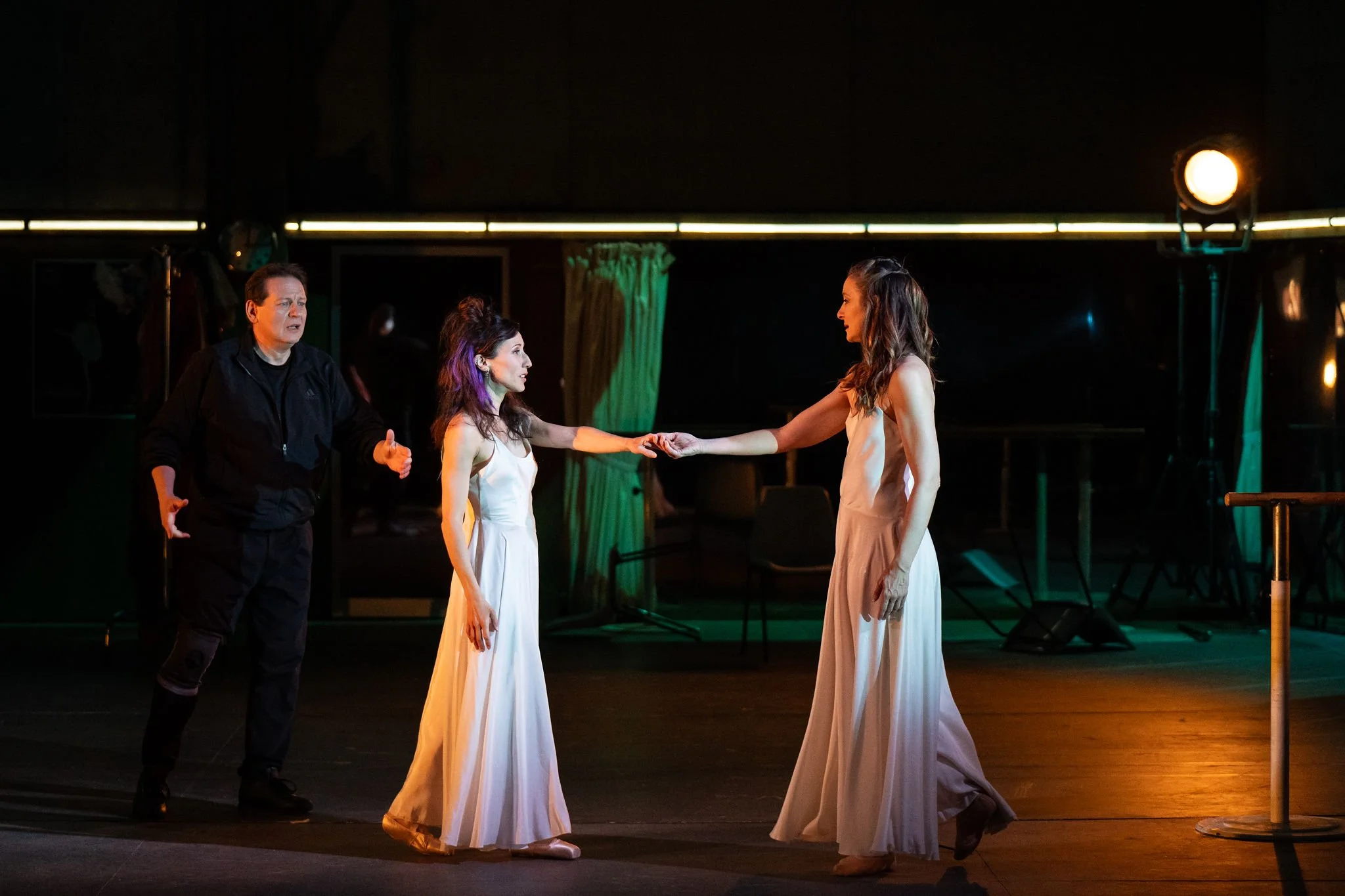 Three women in white dresses on stage, one woman in black clothing appears to be directing or coaching them, in a theatrical setting with dark background and stage lighting.