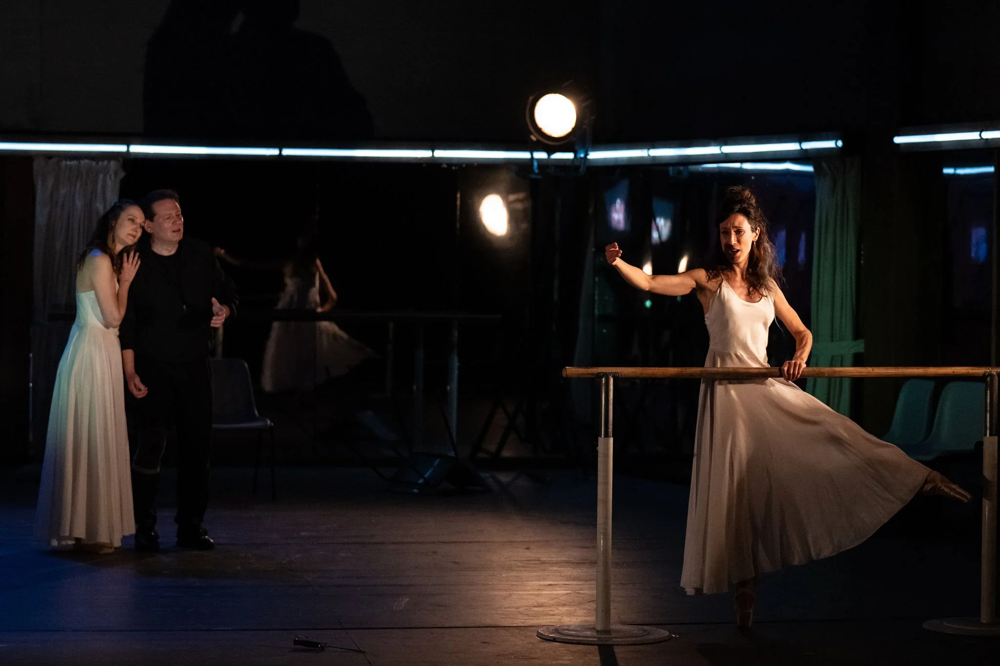 A dance rehearsal or performance with three women in dresses, one woman dancing at barre, and two women watching. The scene is dimly lit with stage lighting overhead.