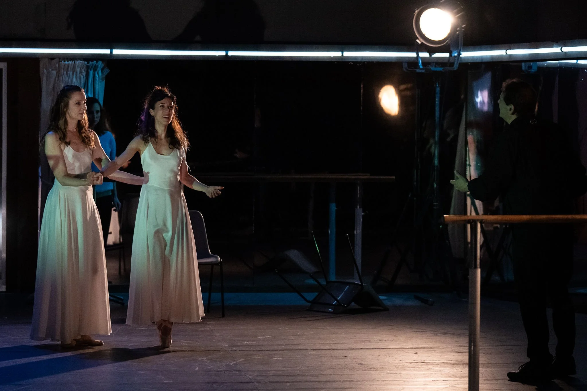 Two women in pastel pink ballet dresses performing on a stage with dark background and stage lighting, while a man in black observes nearby.