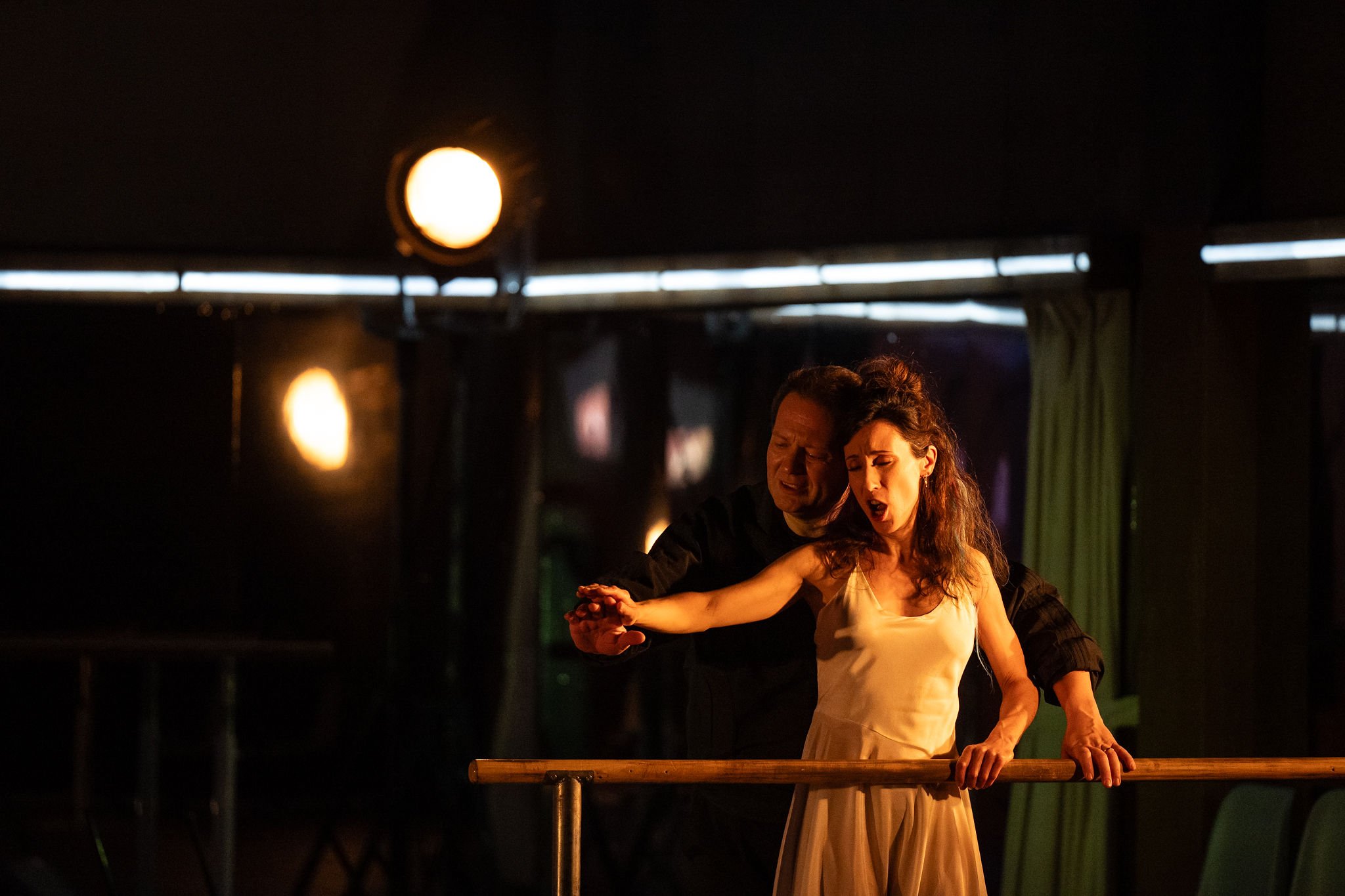 Two performers, a man and a woman, dancing ballet in a dimly lit theater with stage lights overhead.