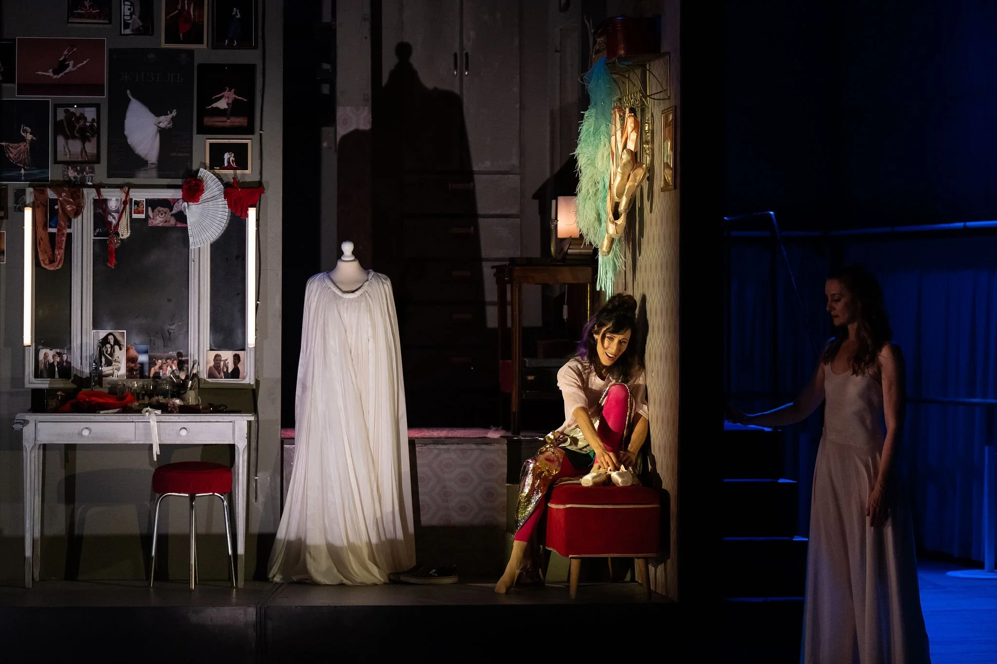 A stage set for a ballet or theater performance with costume accessories, photos, and dance-related decorations, and two women preparing backstage.
