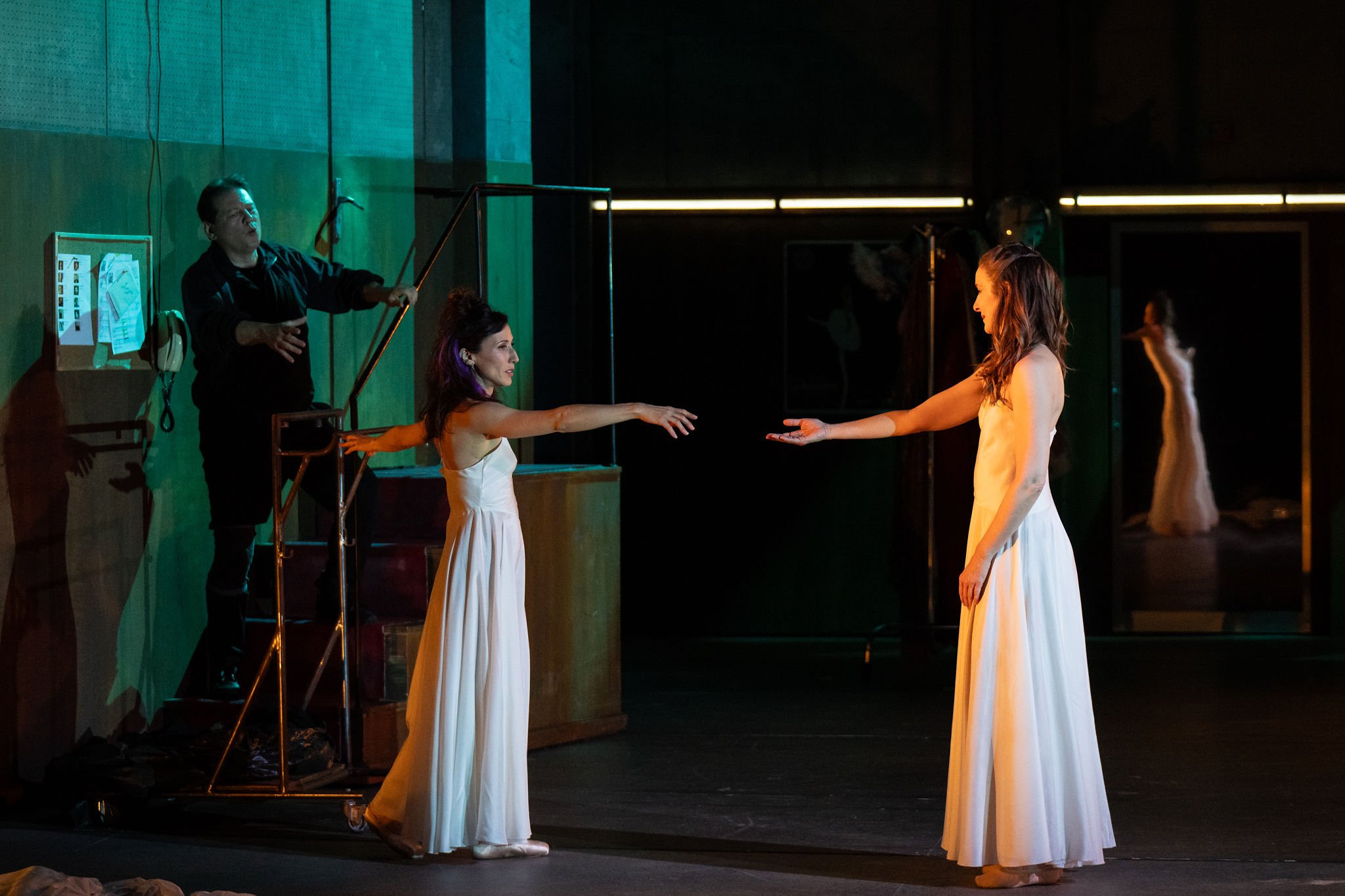 Two ballet dancers in white dresses reaching out to each other, while a man standing on a staircase behind them appears to be adjusting a microphone on a stand, in a dimly lit rehearsal studio.