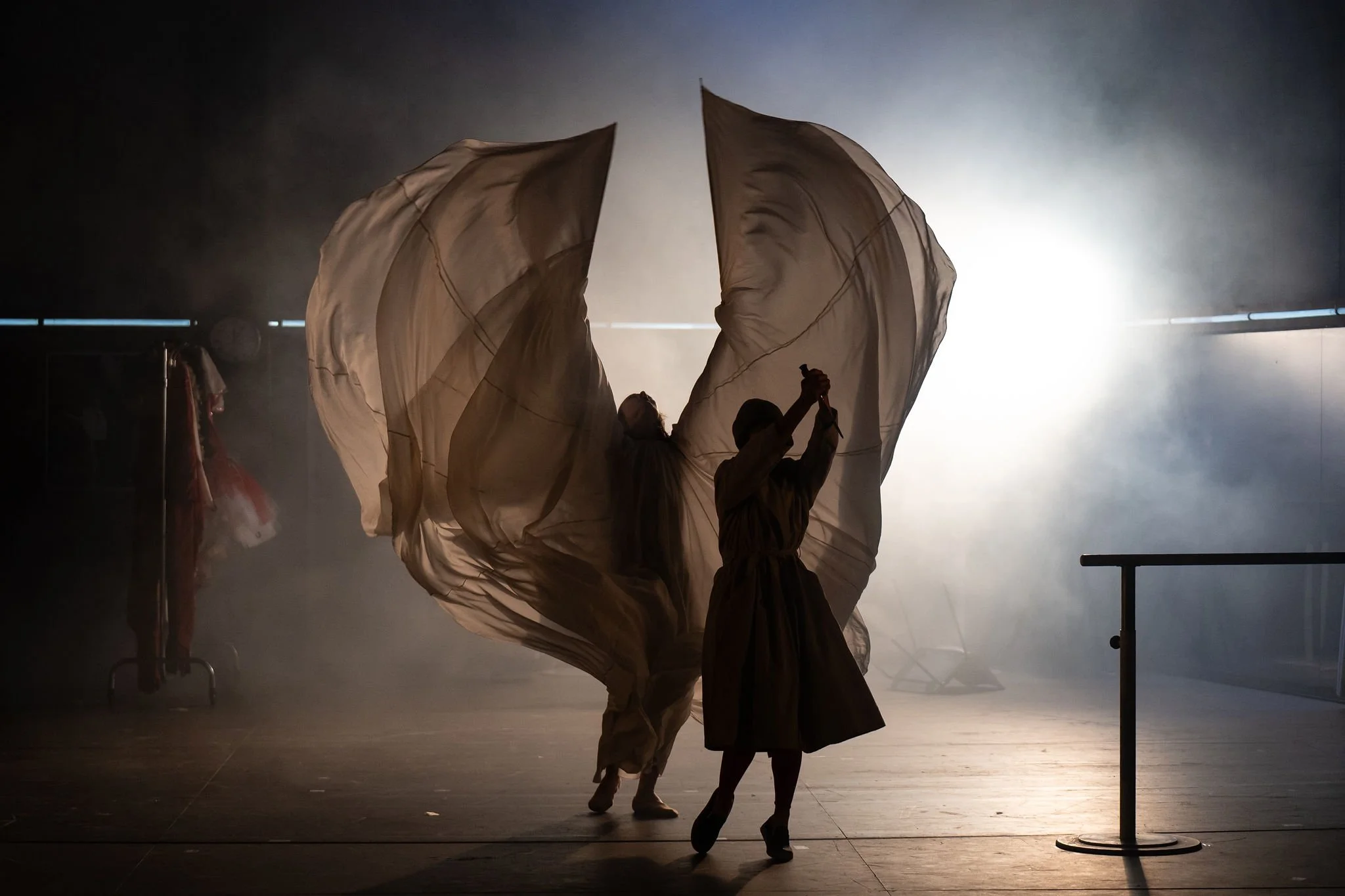 Silhouettes of three performers with large flowing fabric, backlit with bright light, creating dramatic shadows and a misty atmosphere.