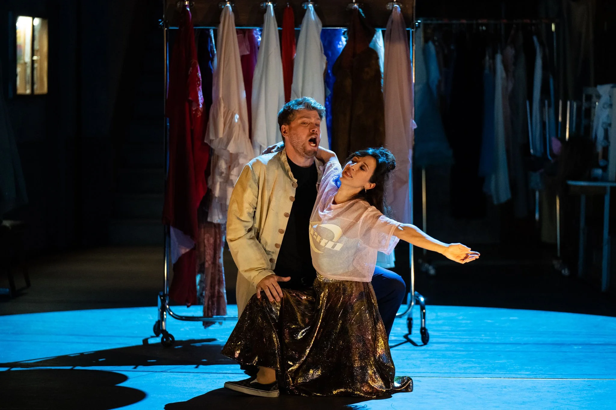 Two actors perform on stage with a clothing rack filled with dresses in the background. The man has light skin, curly hair, and a beard, and is wearing a light-colored jacket and dark shirt. The woman has light skin, long dark hair, and is wearing a 