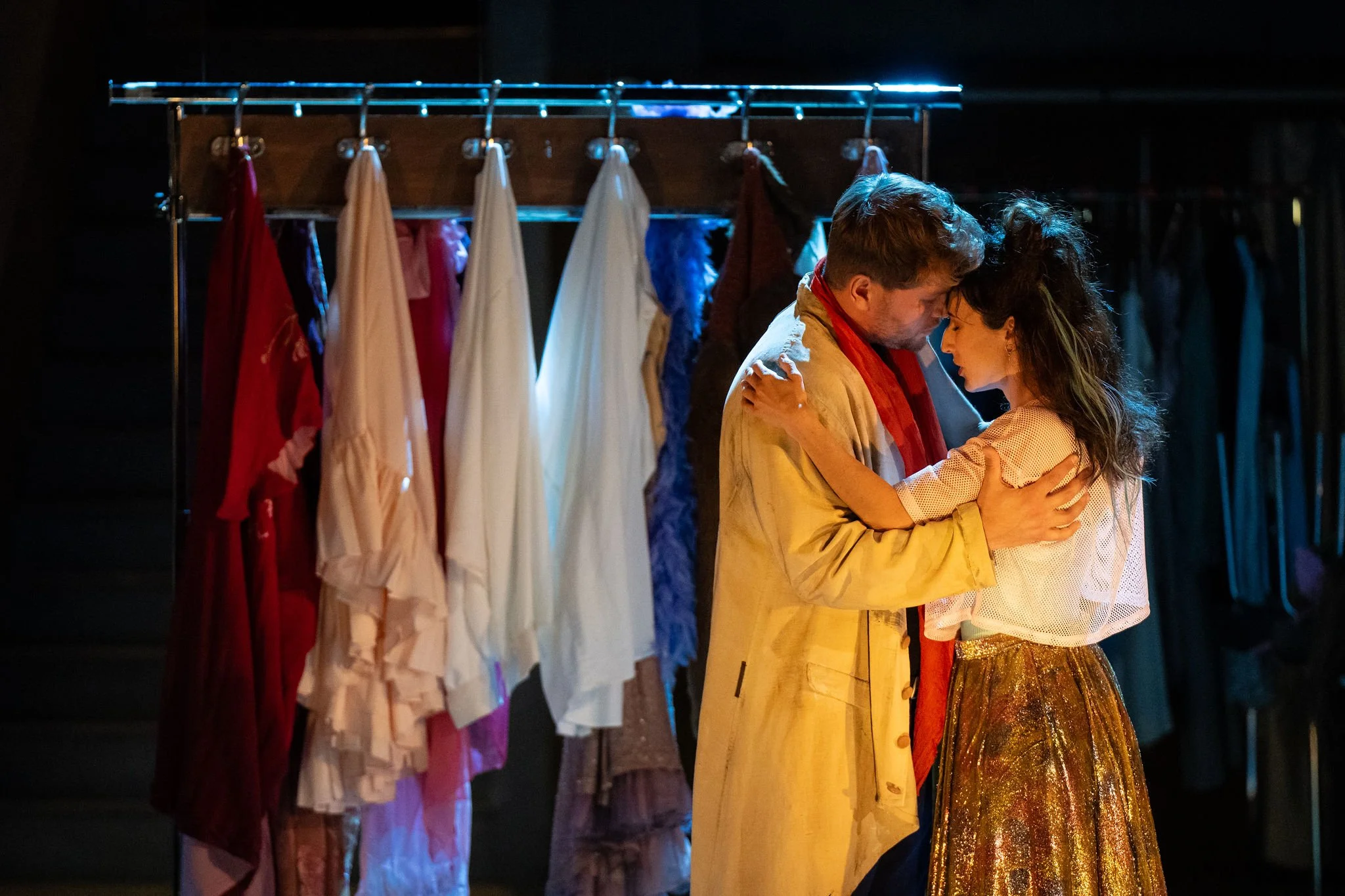 A man and woman embracing backstage at a theater, with costume racks filled with various dresses and garments behind them.