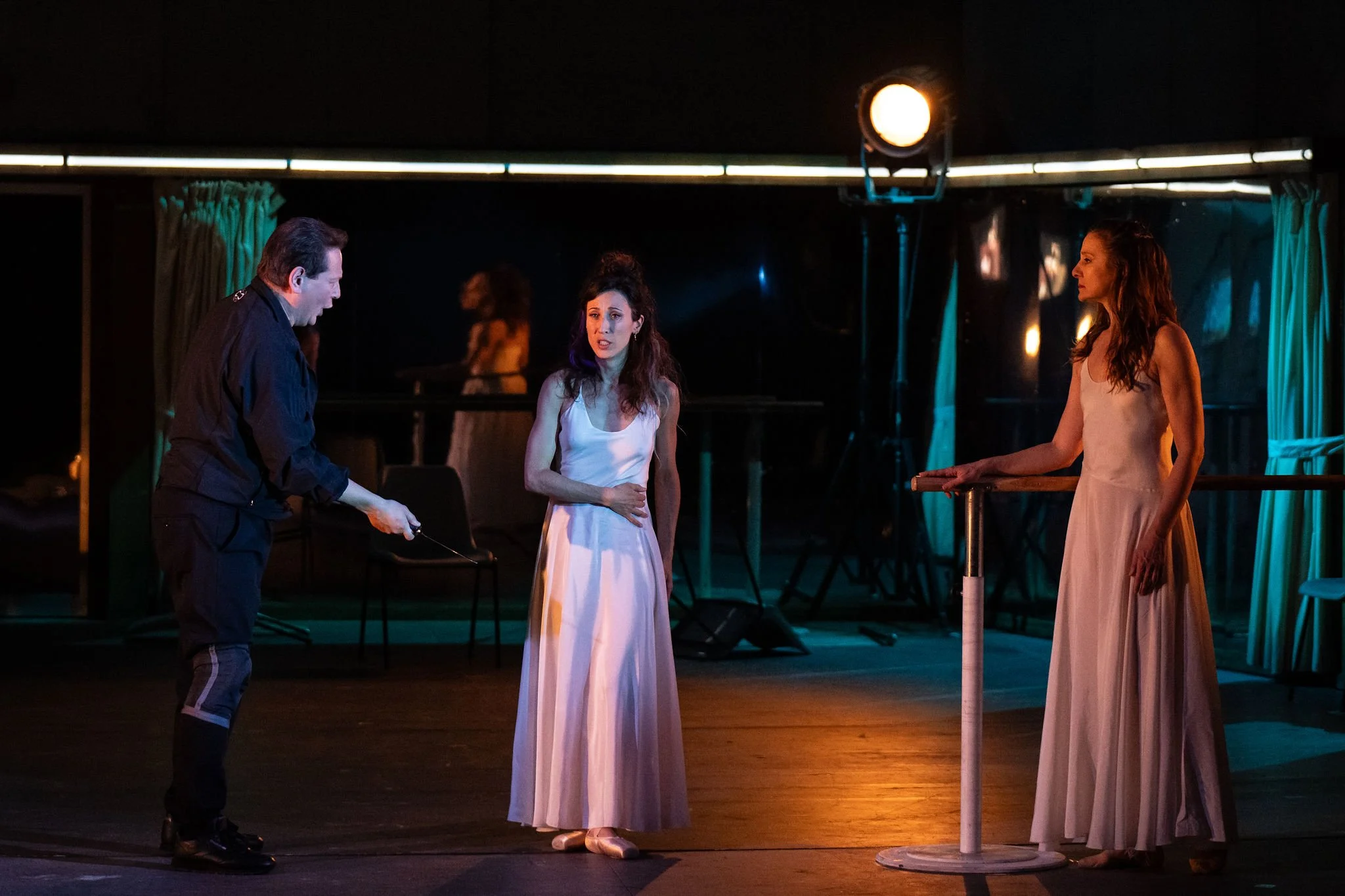 Three women on stage, two in long dresses and one man in dark clothing, engaged in conversation under stage lights, with a reflective background.