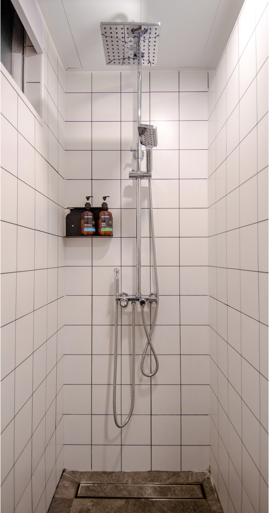 Shower facilities in TeamX Taikoo with white tiled walls, a ceiling-mounted showerhead, a handheld shower wand, and a wall-mounted shelf with two bottles of shampoo and body wash.
