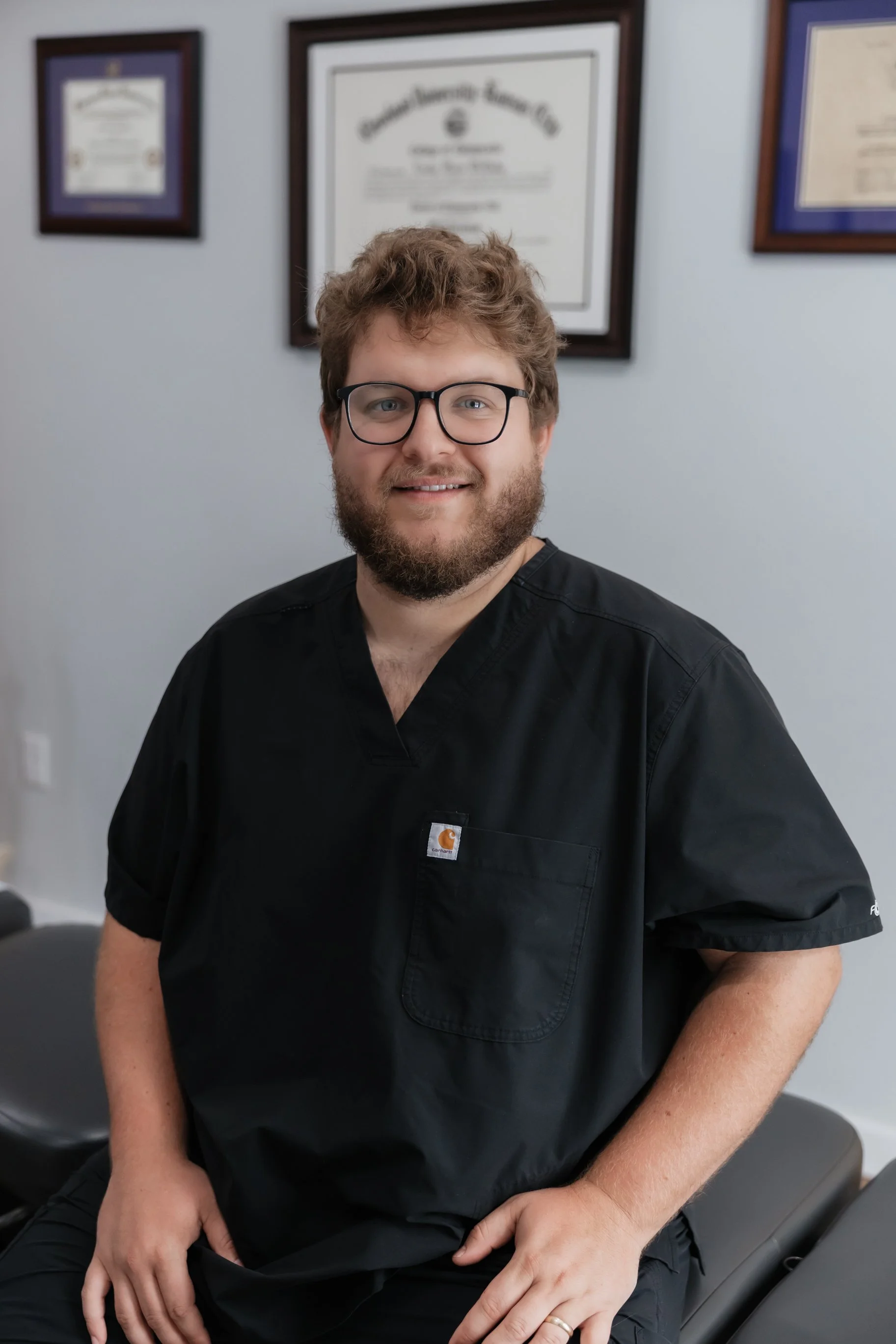 A man with glasses and a beard sitting on a medical examination table, wearing a black medical uniform with a Carhartt logo, in a room with framed certificates on the wall behind him.