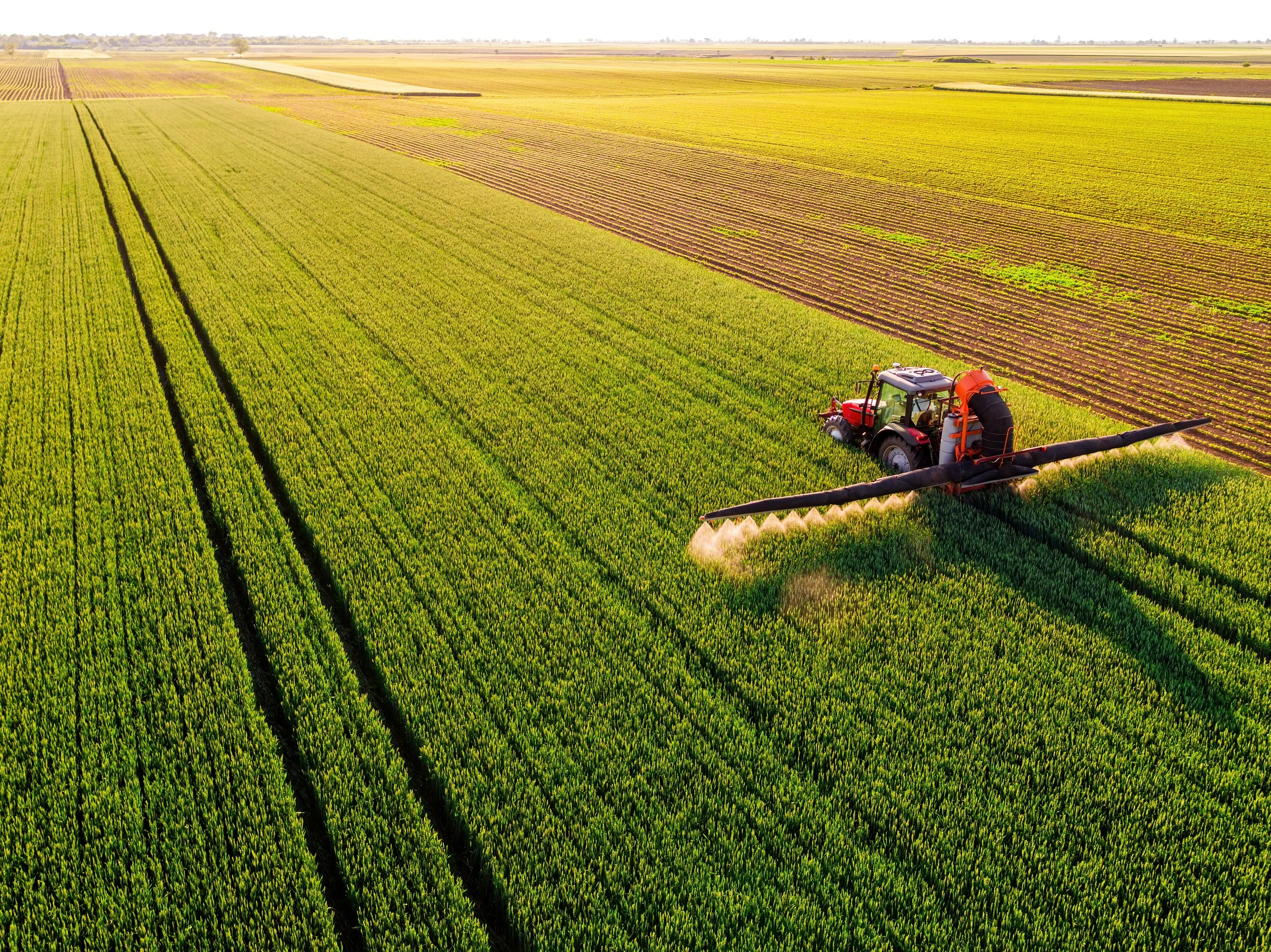 Aerial view of green and brown agricultural fields with a red tractor spraying crops in the bottom right corner.