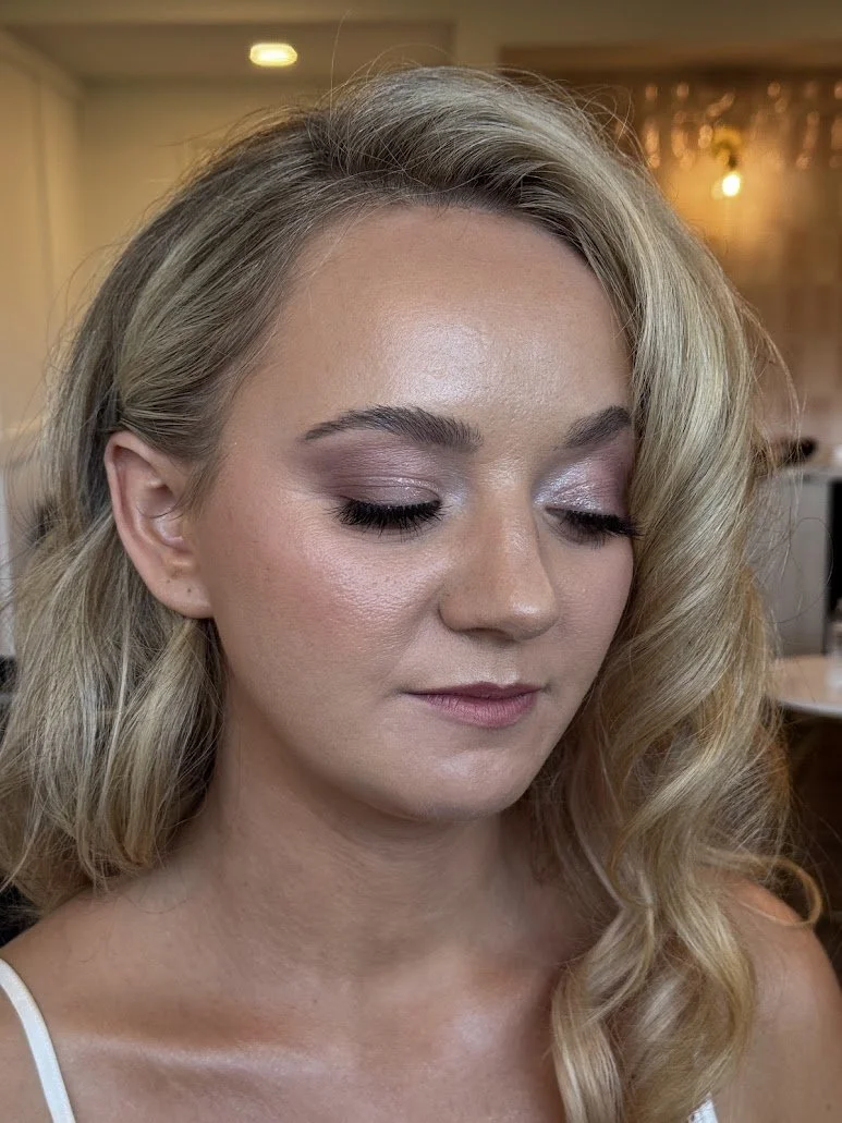 A woman with styled blonde hair and natural makeup with lavender eyeshadow and pink lipstick.