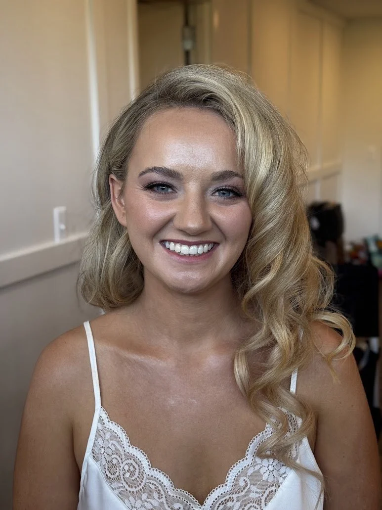 A young woman with blonde, wavy hair and fair skin, smiling at the camera, wearing a white lace-trimmed top, in an indoor setting with beige walls and a blurred background.