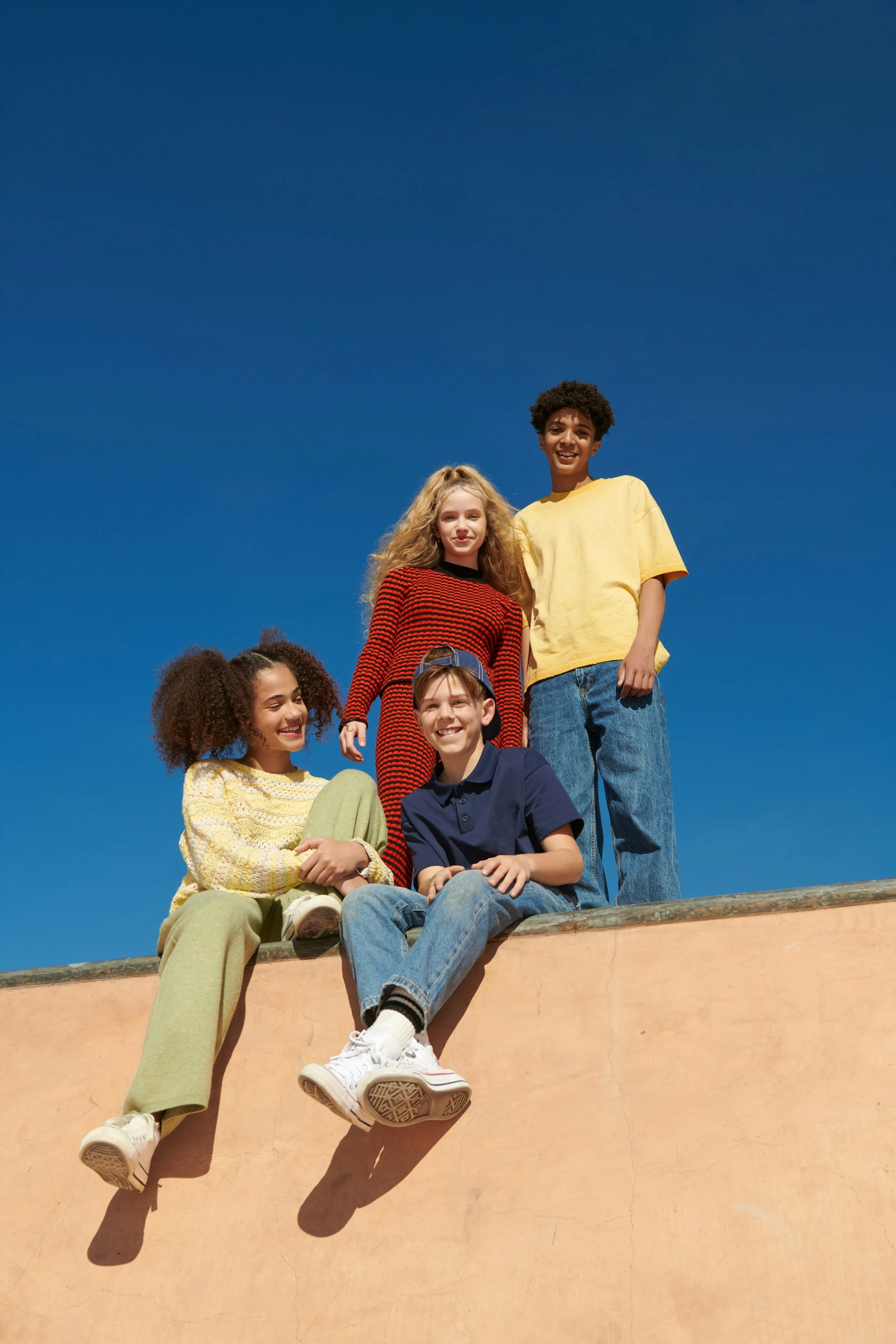 Group of five young friends, three girls and two boys, smiling and relaxed outdoors on a sunny day, sitting and standing on a ledge with a clear blue sky in the background.