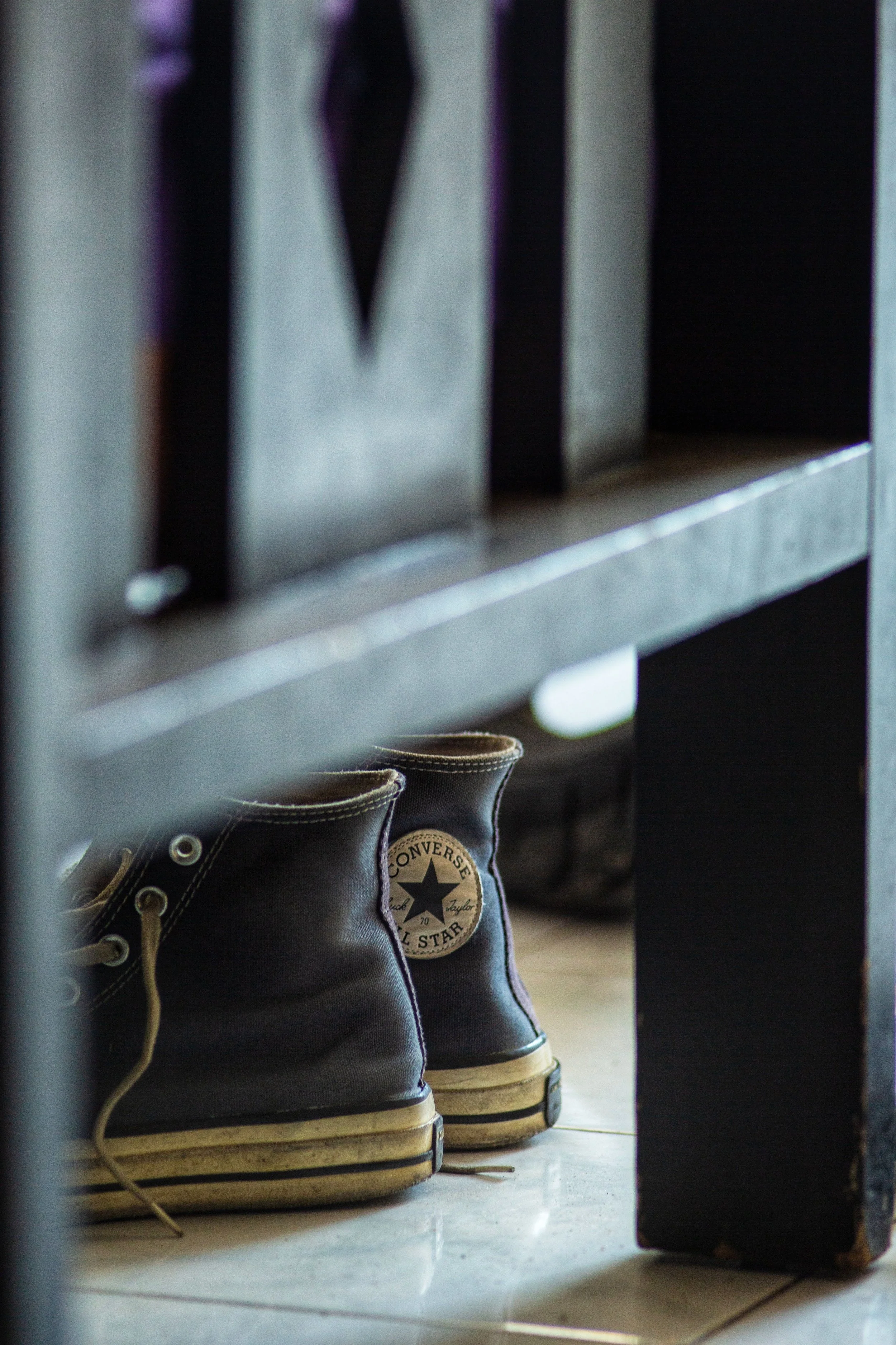 A pair of black Converse All Star sneakers with beige soles and white laces on a tiled floor, positioned in front of a metal shelf or bench.