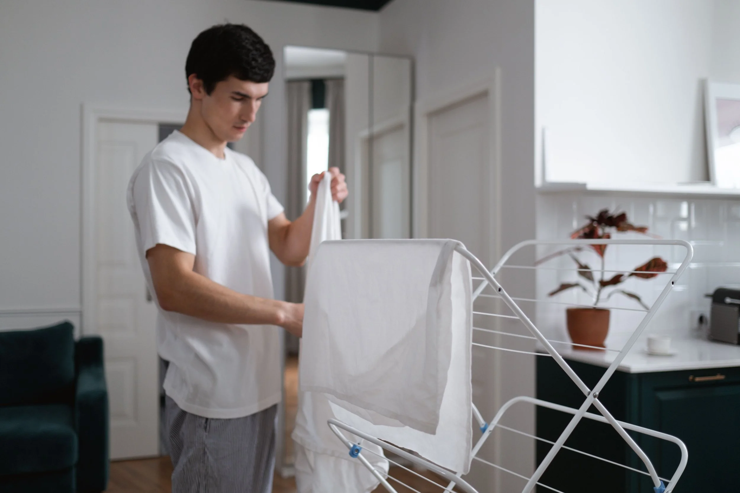 A young man with dark hair, wearing a white t-shirt and striped pajama pants, is folding laundry in a bright, modern room. There is a white drying rack in front of him and a potted plant on a table in the background.