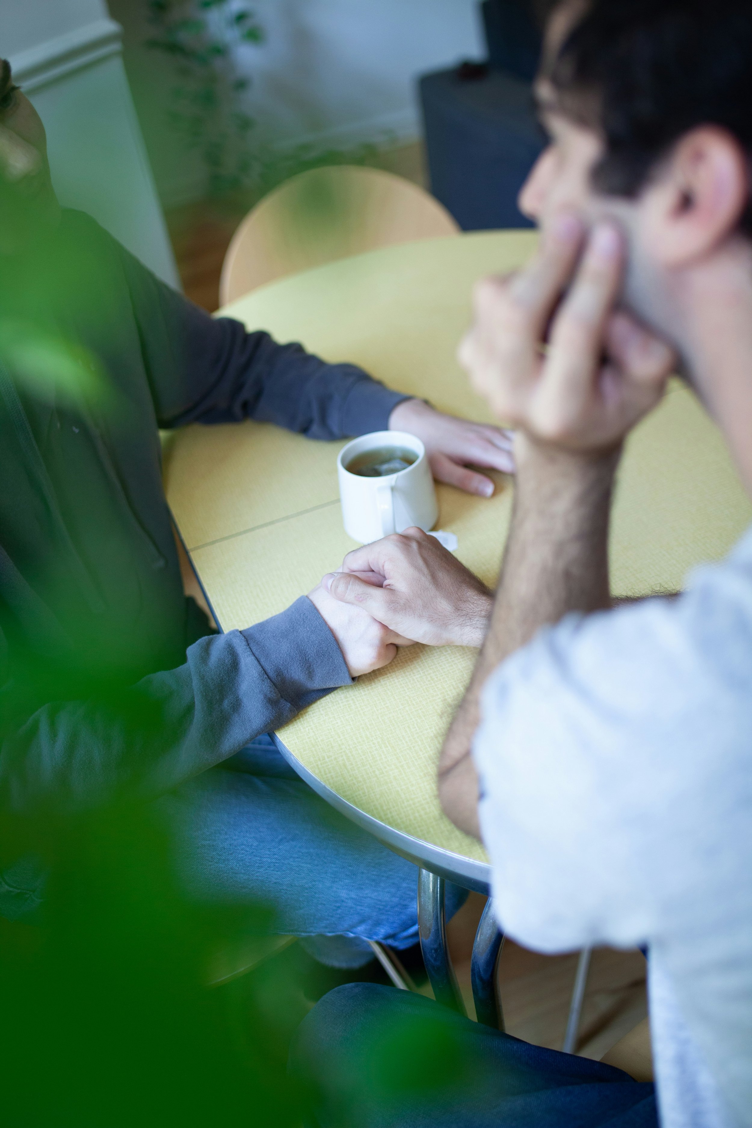 Two men sitting at a yellow table, having a discussion. One man is holding a white coffee mug, while the other is resting his chin on his hand. The scene is indoors with some visible furniture and plants.
