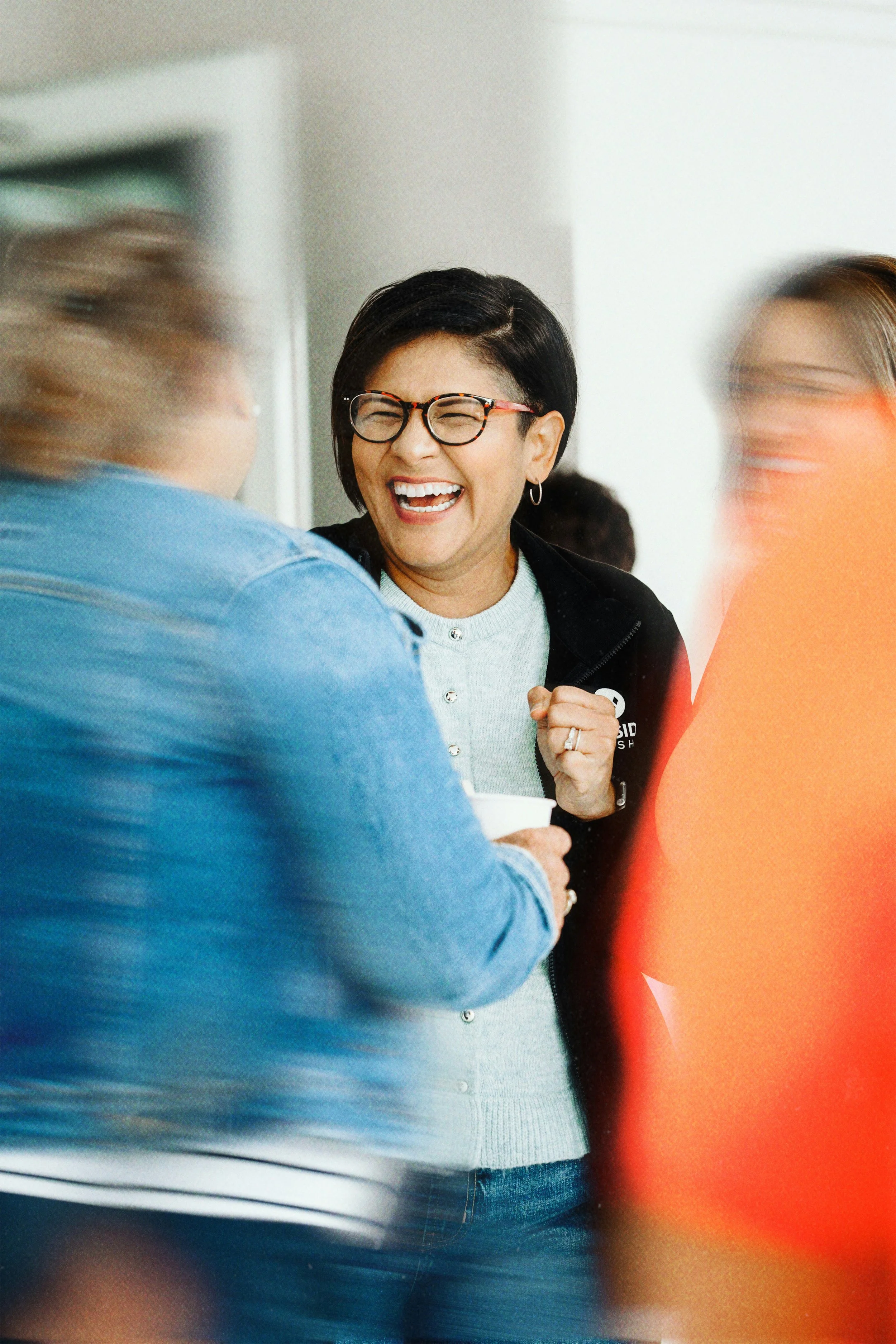 A woman with short black hair, glasses, and hoop earrings laughing and talking to two other blurred women in a social setting.