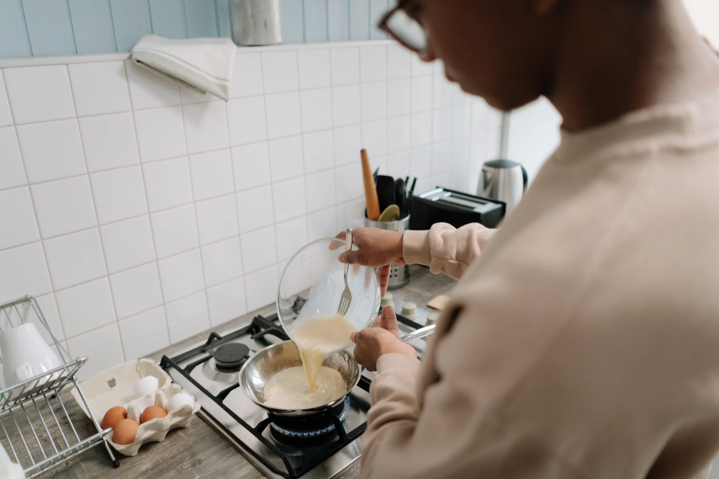 A person is cooking on a stovetop, pouring batter from a bowl into a pan. The kitchen has a white tiled wall, eggs in an egg carton on the counter, and kitchen utensils nearby.