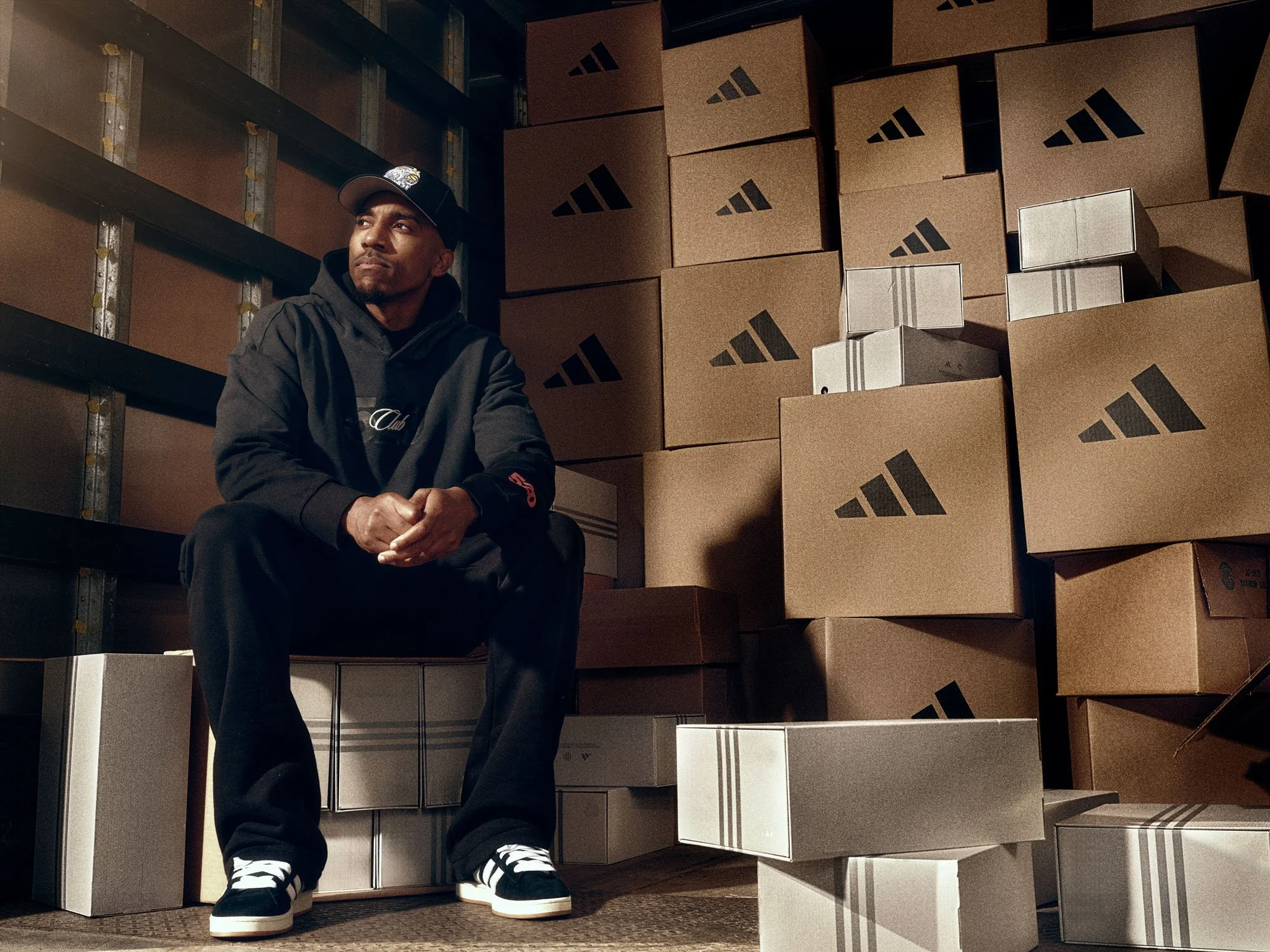 A man sitting in a warehouse surrounded by stacked cardboard boxes with the Adidas logo. He is wearing a black hoodie, black pants, and black sneakers with white soles and laces, and a cap. Jeff Teague club 520 photographed by Jay Goldz