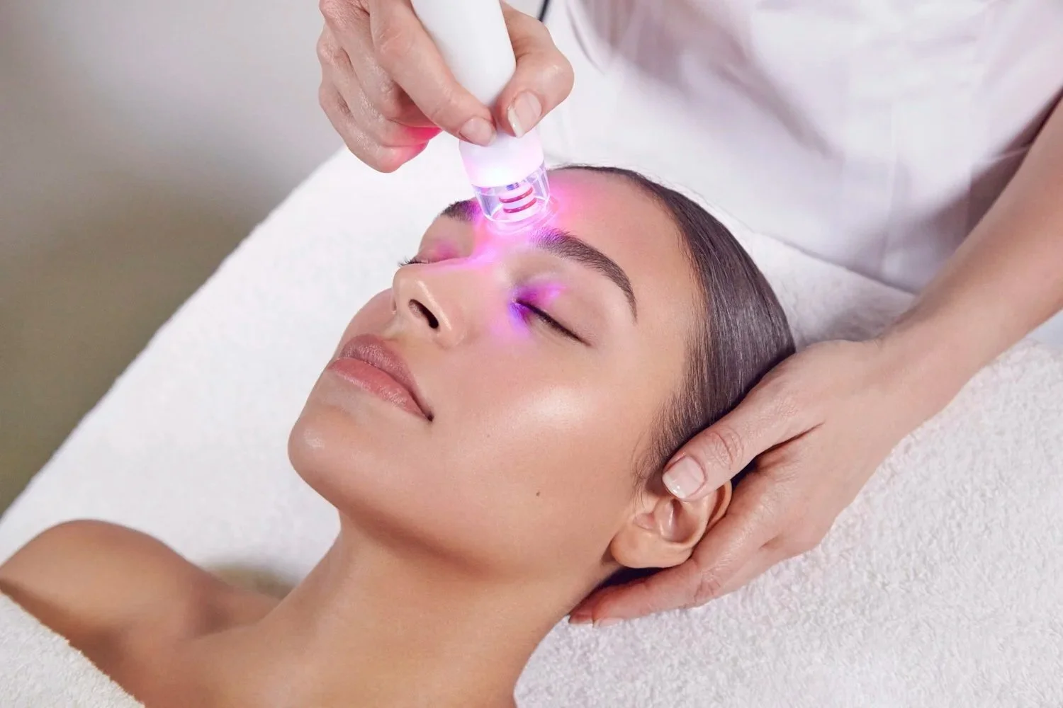 A woman receiving a facial treatment with a handheld device emitting pink and purple light, lying on a massage table with her eyes closed.
