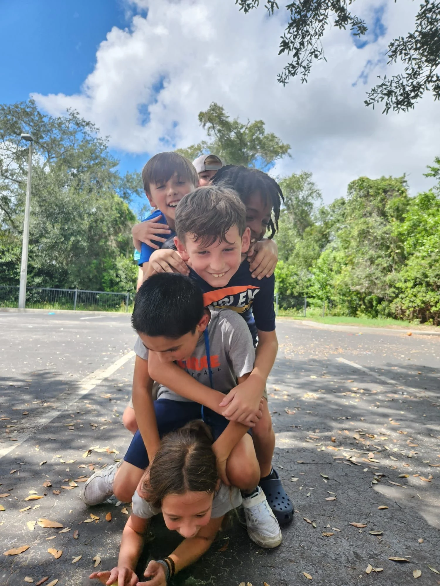 Six children play together outdoors, forming a human stack on an asphalt surface with scattered leaves, surrounded by trees and blue skies with clouds.