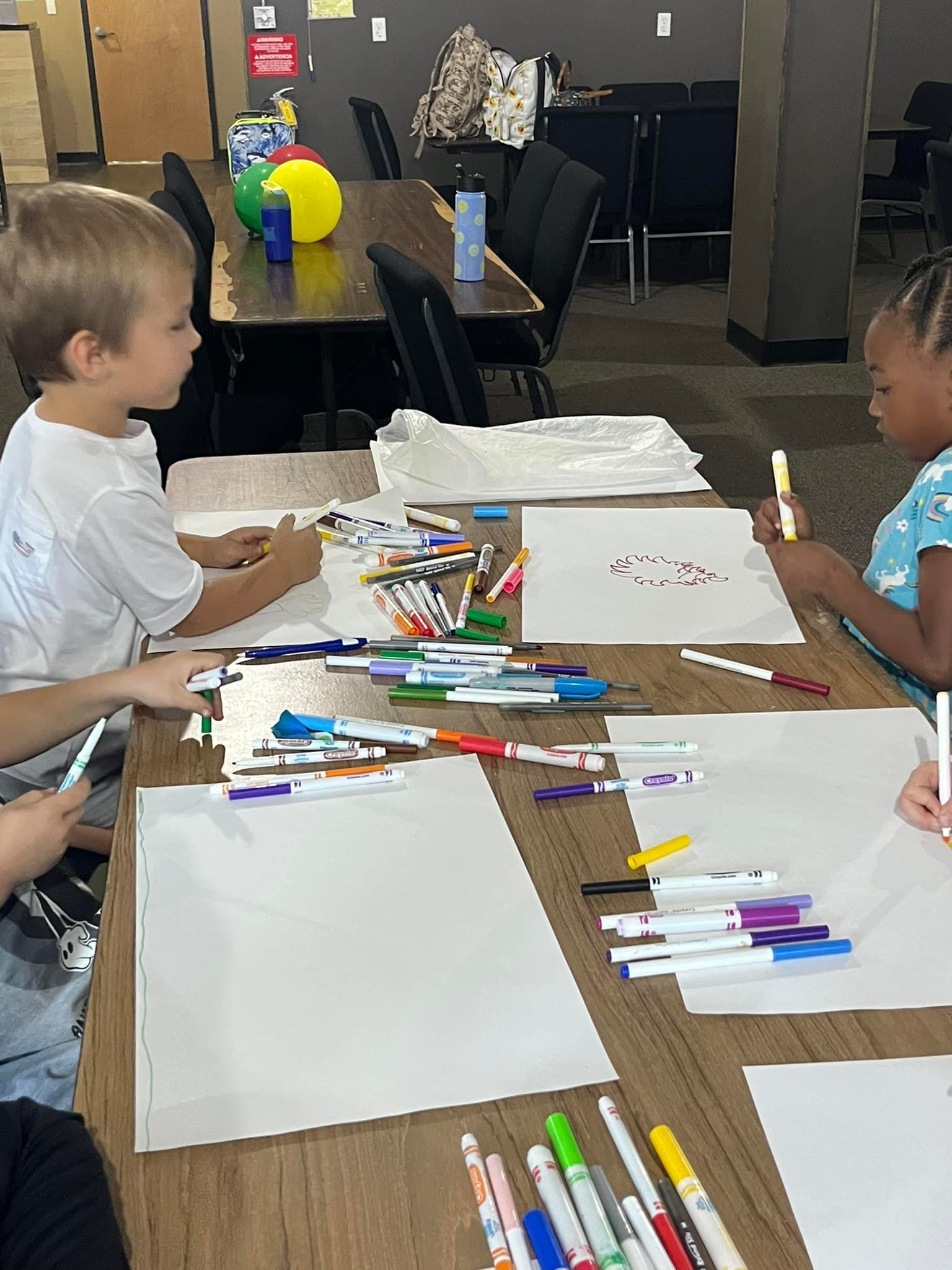 Children sitting around a table drawing and coloring on large sheets of paper with markers in a classroom or activity room.