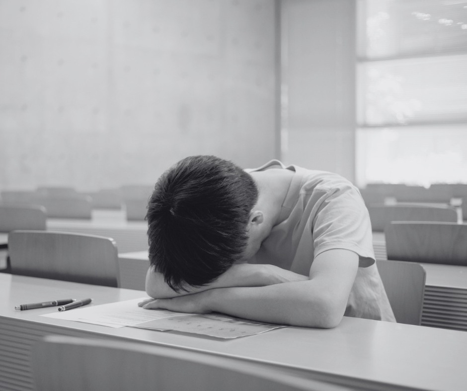 A student sits at a desk in an empty classroom with his head resting on his arm, appearing exhausted or stressed, with pens and papers on the desk.