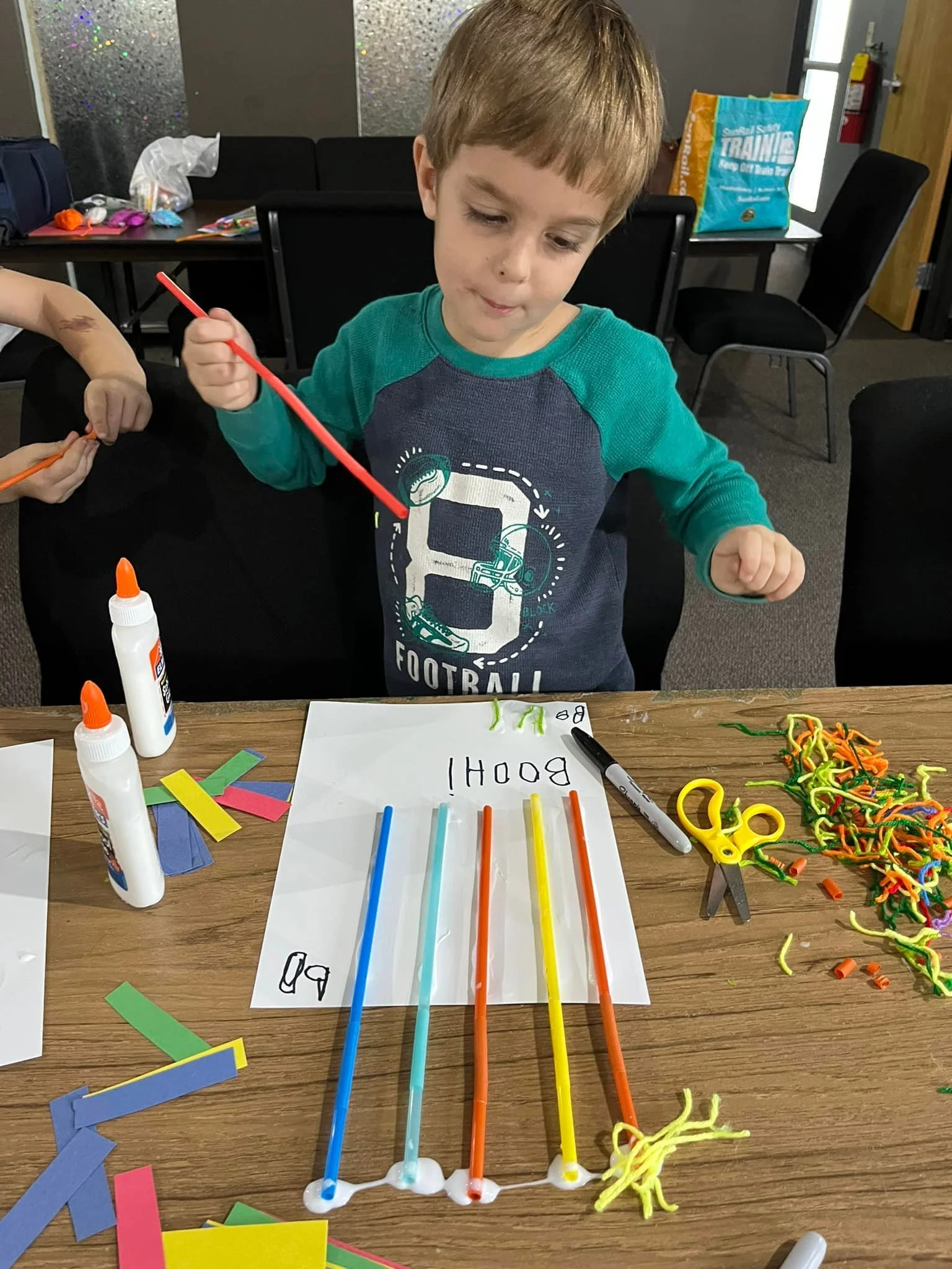 A young boy with light brown hair wearing a green and dark blue long sleeve shirt creating a science experiment with colorful spaghetti and glue at a table, with some paper and scissors nearby.