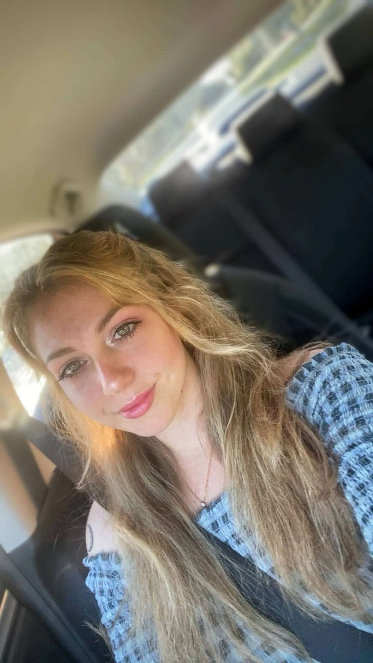 A young woman with long blonde hair and blue eyes taking a selfie in a car, smiling slightly, with sunlight illuminating her face.