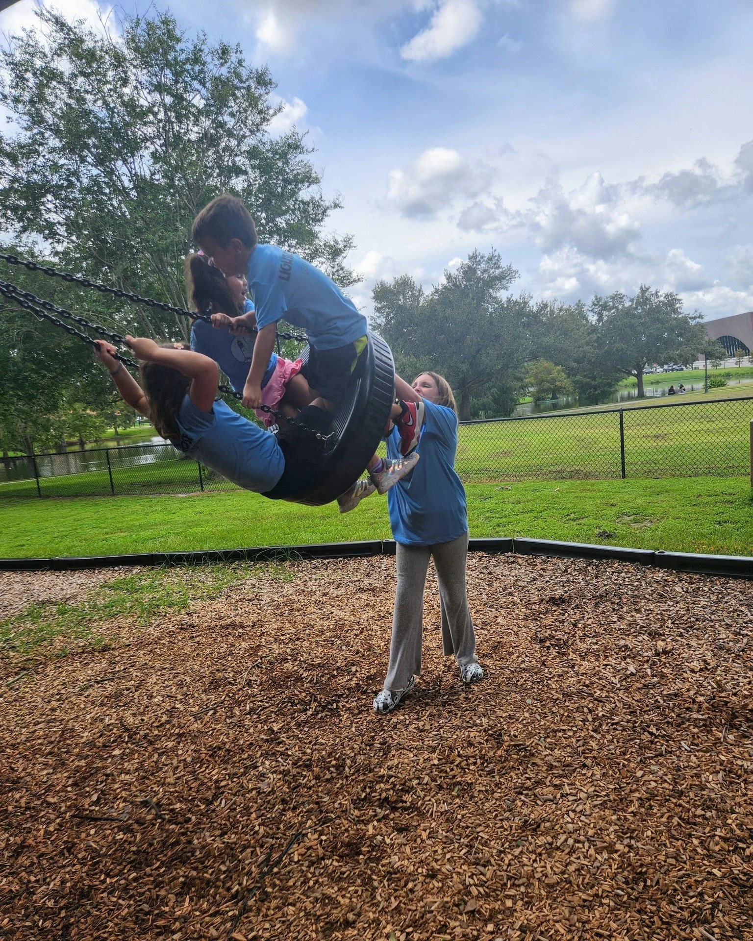 Children playing on a tire swing at a park, with adults assisting, surrounded by grass, trees, a fence, and a blue sky with clouds.