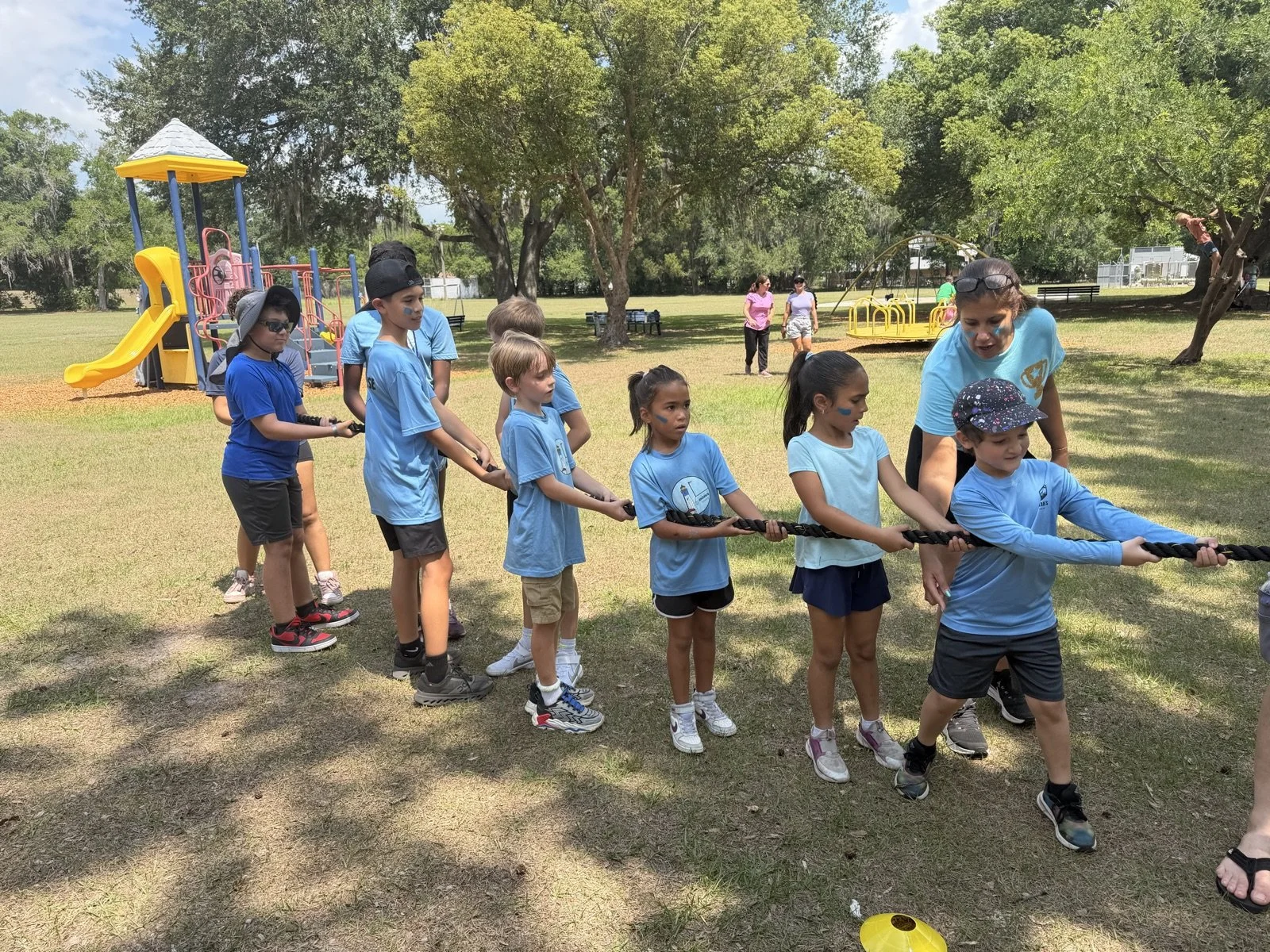 Children participating in a tug-of-war game at a park on a sunny day, with playground equipment and trees in the background.
