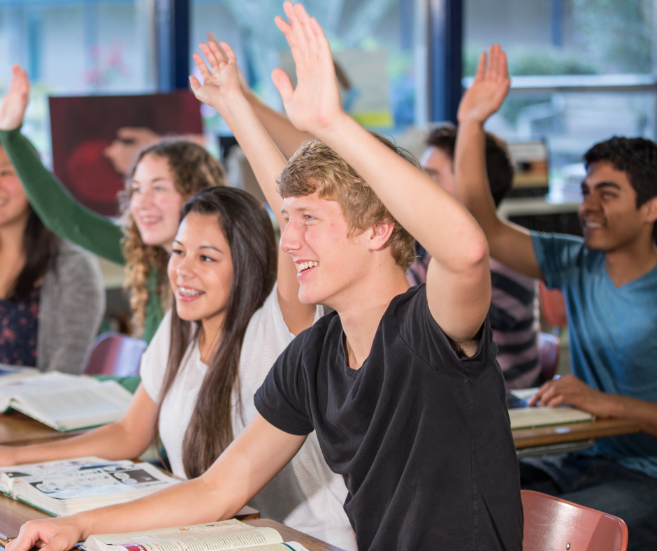 Students raising hands in classroom, smiling and engaging in class activity.