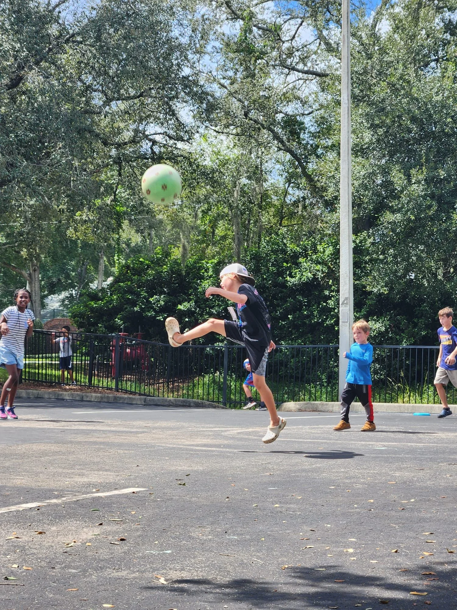 Kids playing soccer on a court on a sunny day, with a boy jumping and kicking a green ball, surrounded by other children watching.