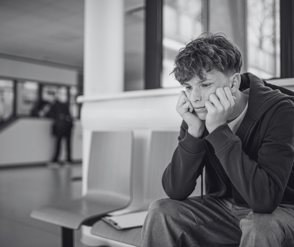 A young boy sitting on a bench at an indoor waiting area, resting his head on his hands with a contemplative expression, while a blurred figure stands in the background near a window.